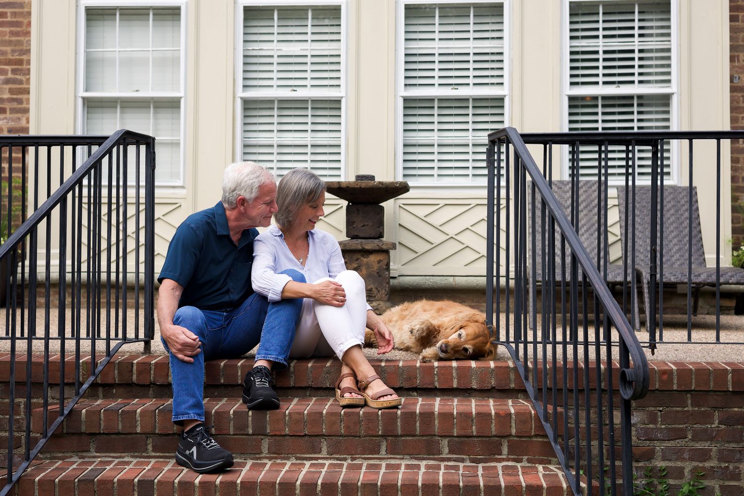 Elderly couple sitting on brick steps sharing a tender moment with a golden retriever dog resting beside them.