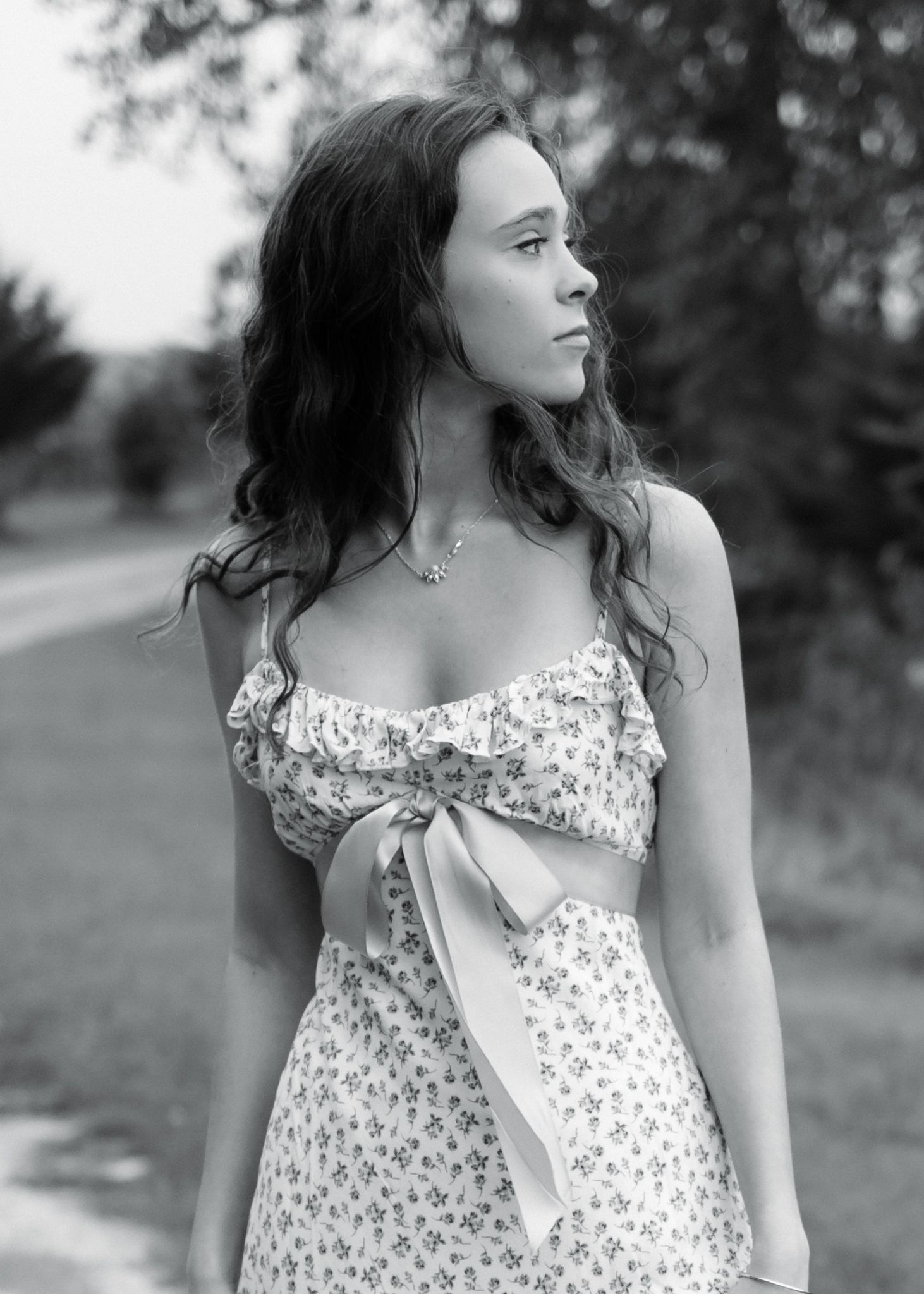 Black and white portrait of a young woman in a floral crop top with bow, standing outdoors on a country road.