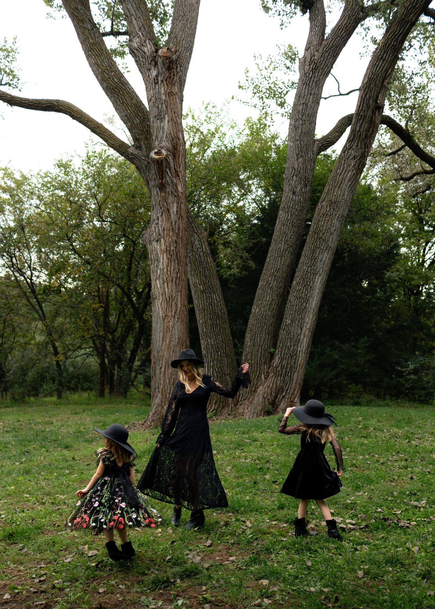 Three figures in black witch costumes and hats dance near large trees in a forest setting.