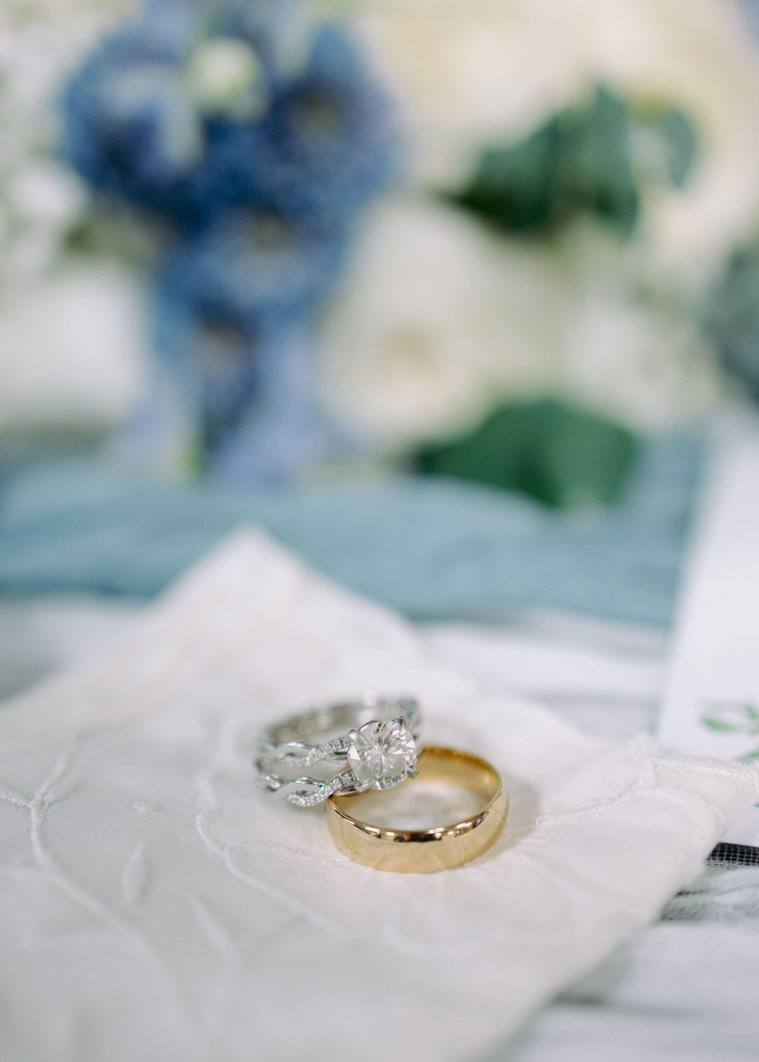 Diamond engagement ring and gold wedding band resting on a white linen pouch with soft blue and white wedding flowers blurred behind.