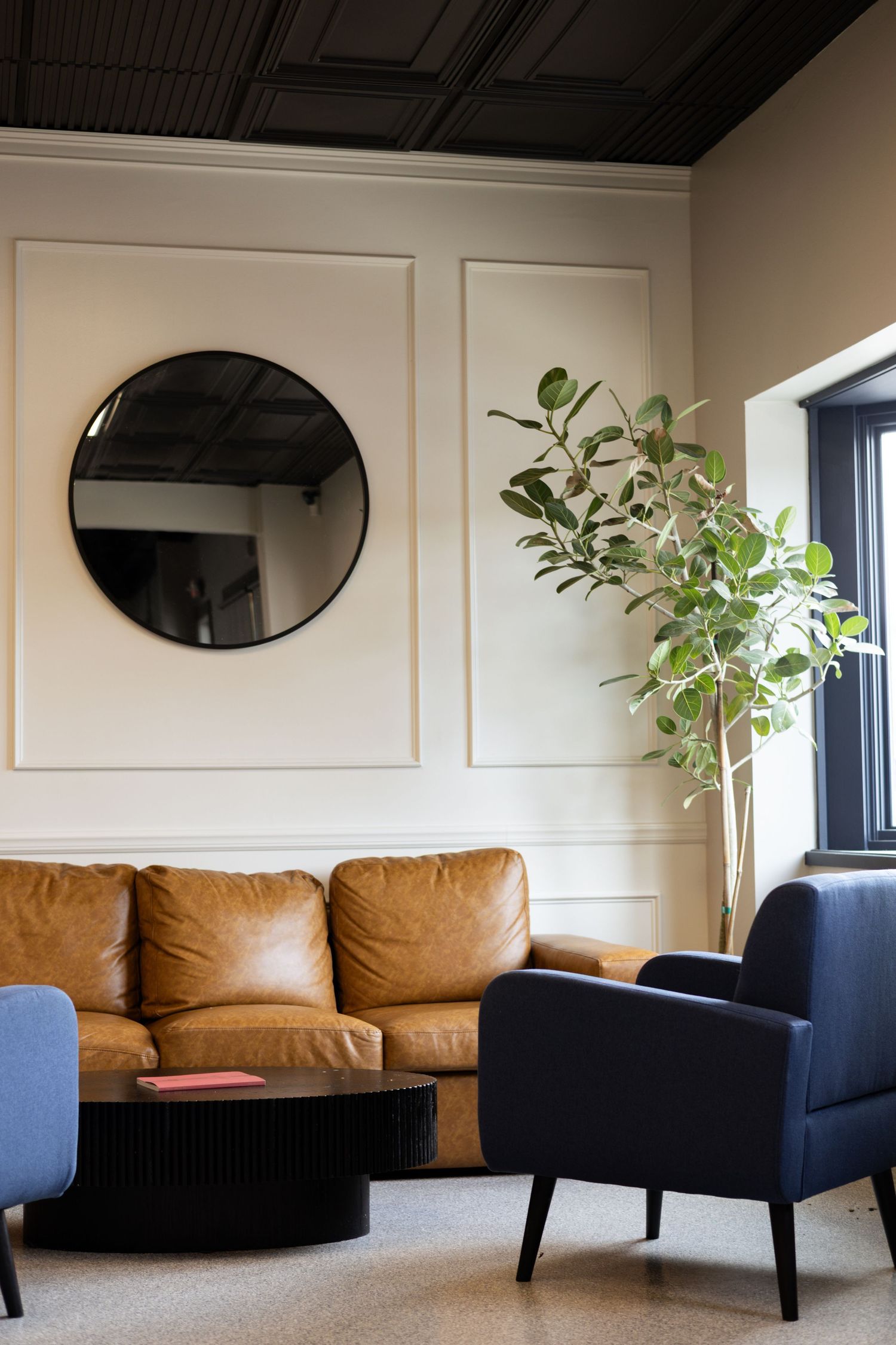 Modern living room with tan leather sofa, navy armchair, round black mirror, and indoor plant near black-framed window.