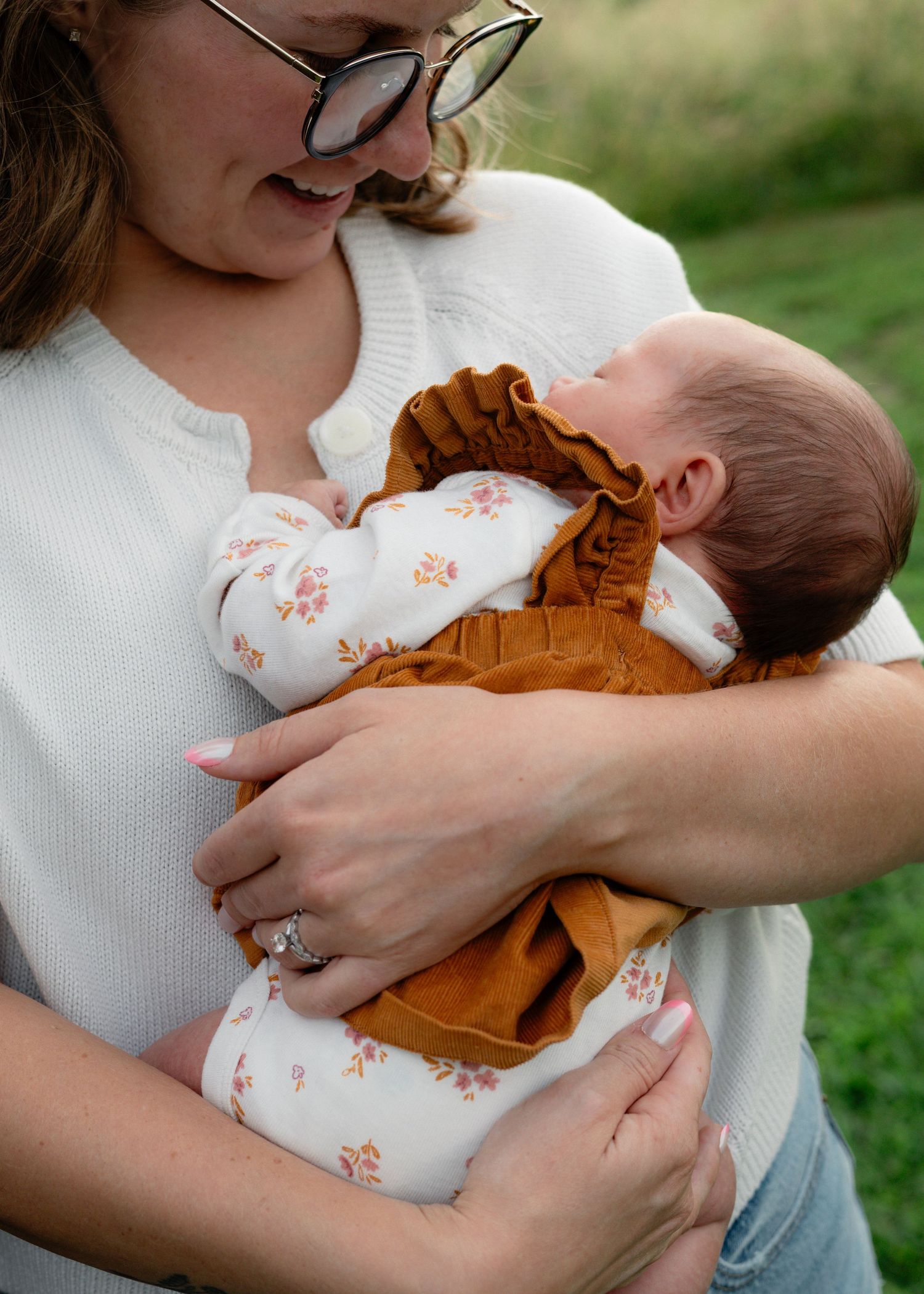 A mother in a white sweater tenderly holds her newborn baby dressed in a floral outfit outdoors.