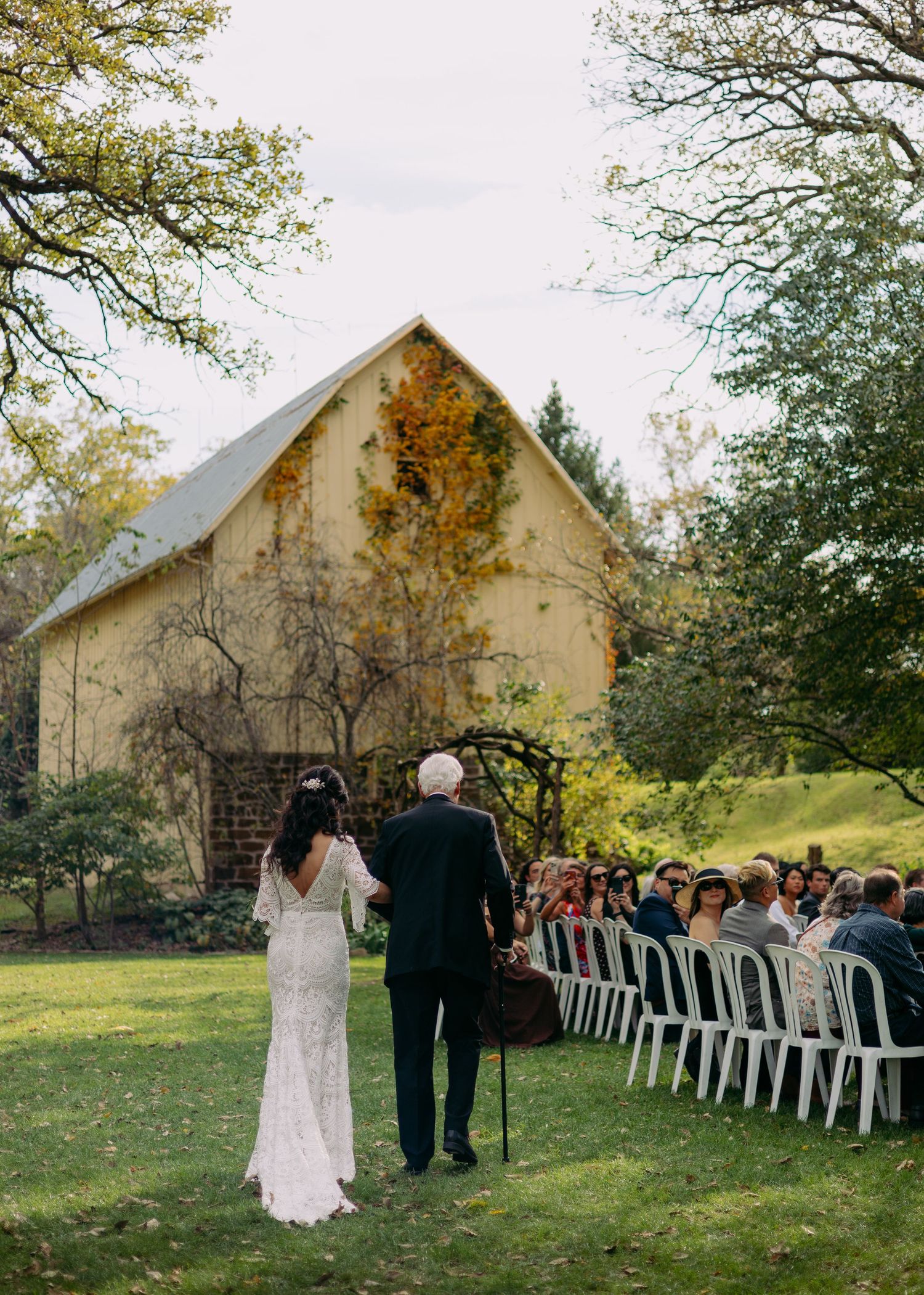 Bride in a lace gown walks down the aisle on a grassy lawn toward seated wedding guests near an ivy-covered rustic barn.