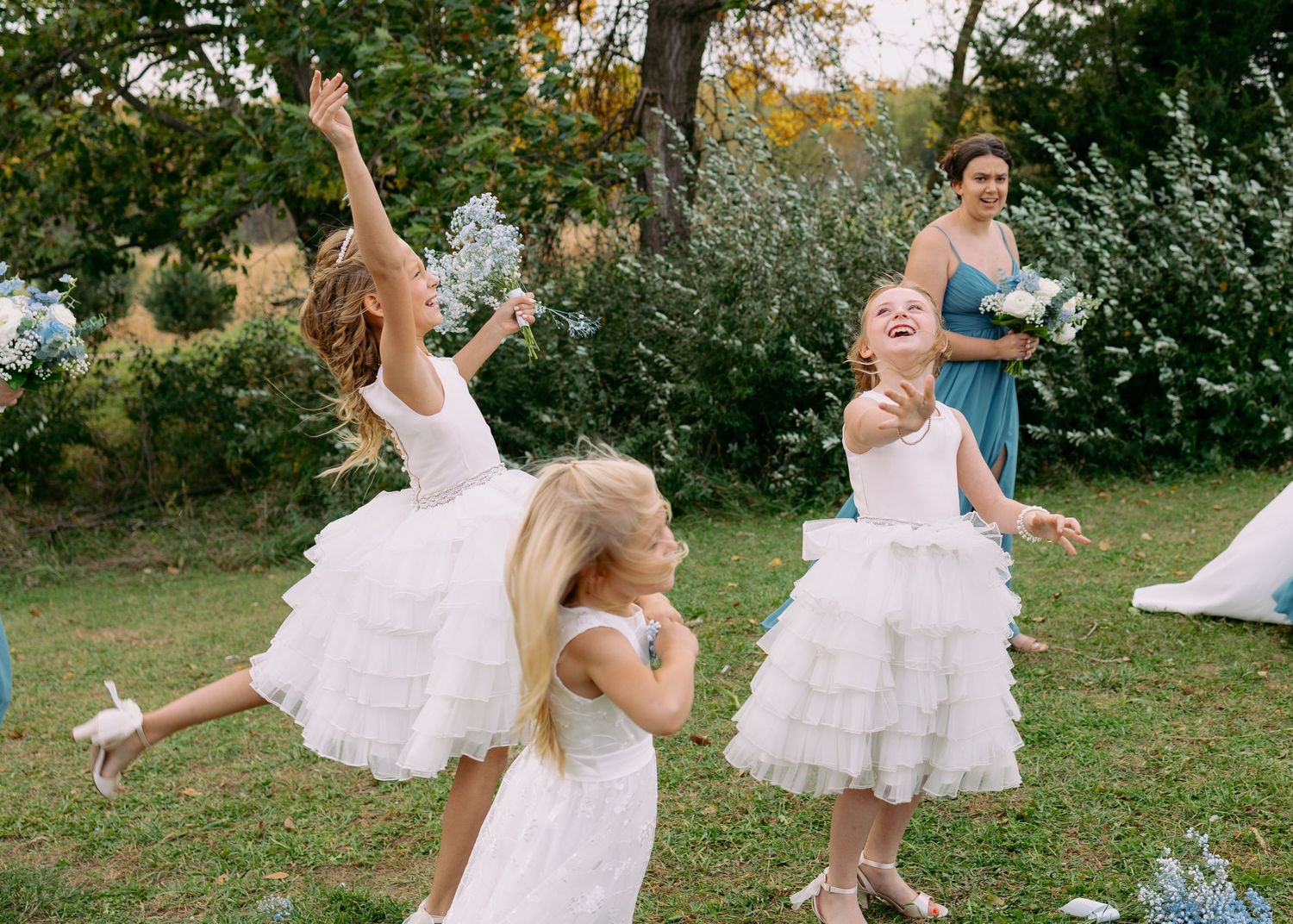 Three flower girls in white tulle dresses dance joyfully on a sunny lawn while a bridesmaid in blue holds a bouquet nearby.