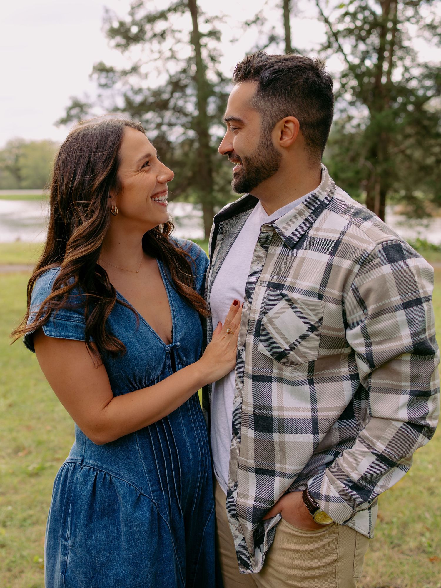 Smiling couple embracing outdoors, a woman in a denim dress gazing at a bearded man in a plaid shirt, with a lake and trees behind.