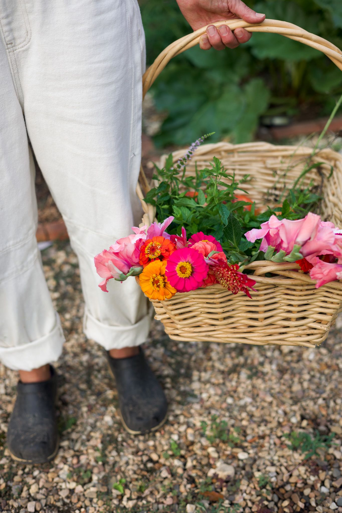 Person in white overalls holding a wicker basket of bright mixed flowers on a gravel path.