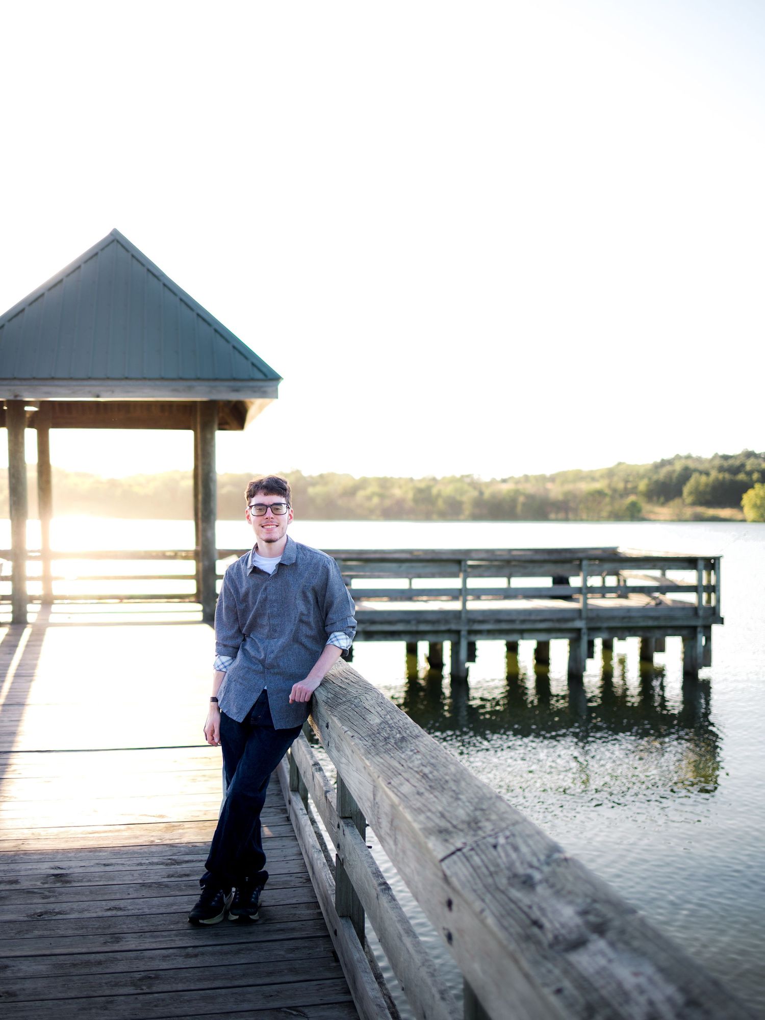 Young man in sunglasses poses on a wooden dock by a calm lake at golden hour sunset.