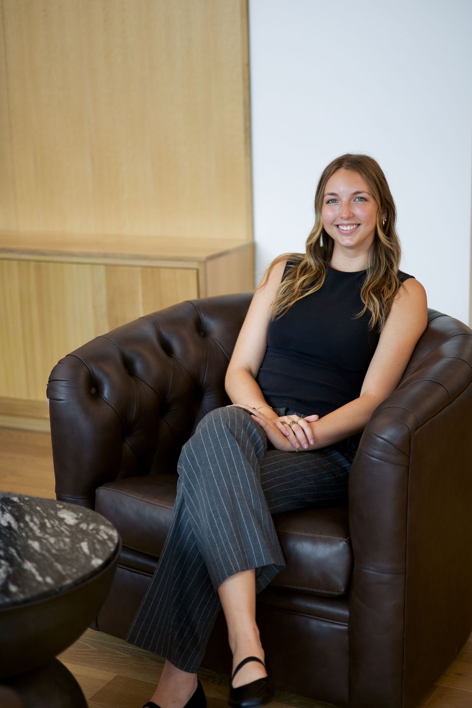 Smiling young professional woman sitting in a tufted leather armchair in a modern office setting.