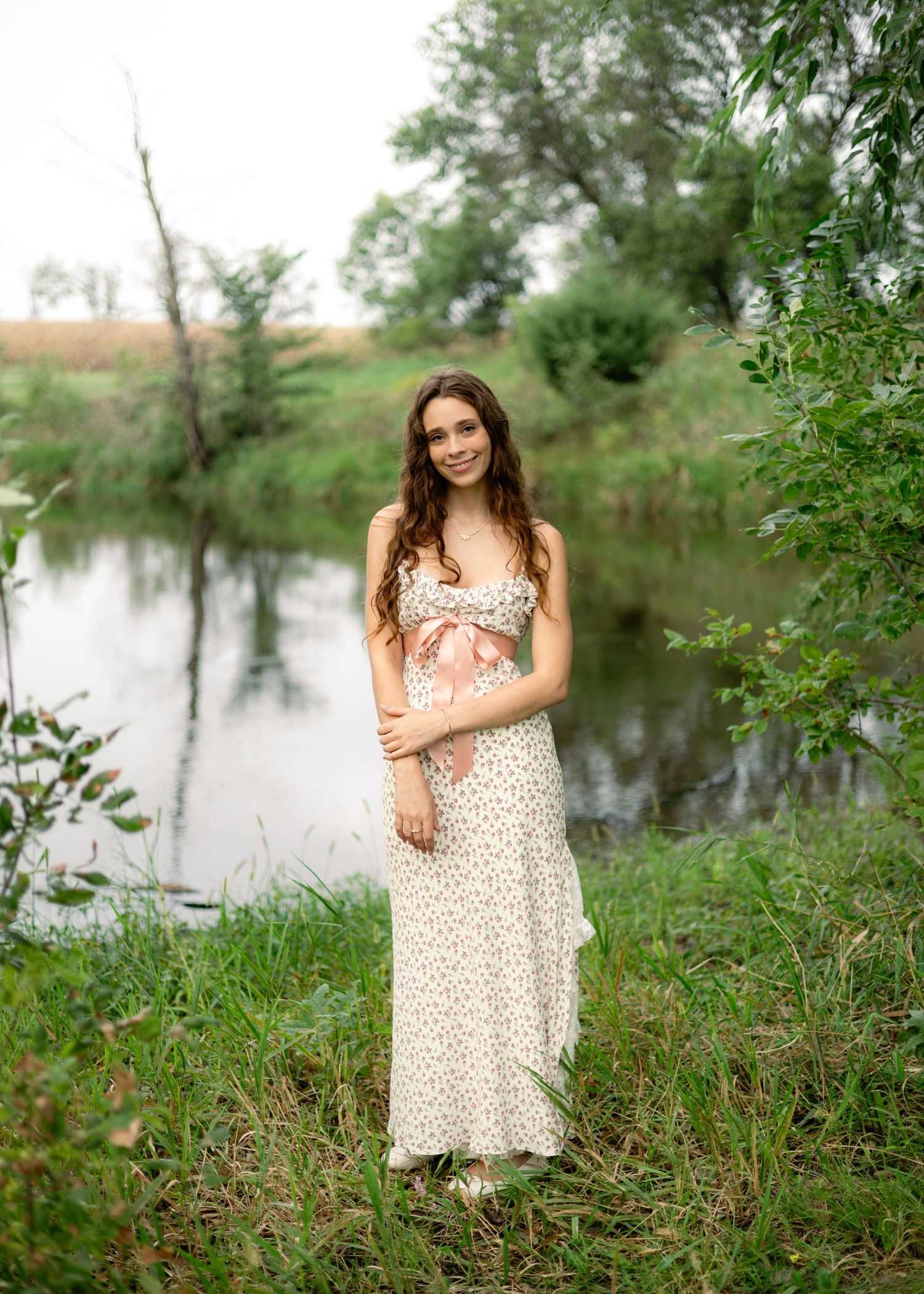 Young woman in floral maxi dress posing by a serene creek surrounded by lush green trees and grass.