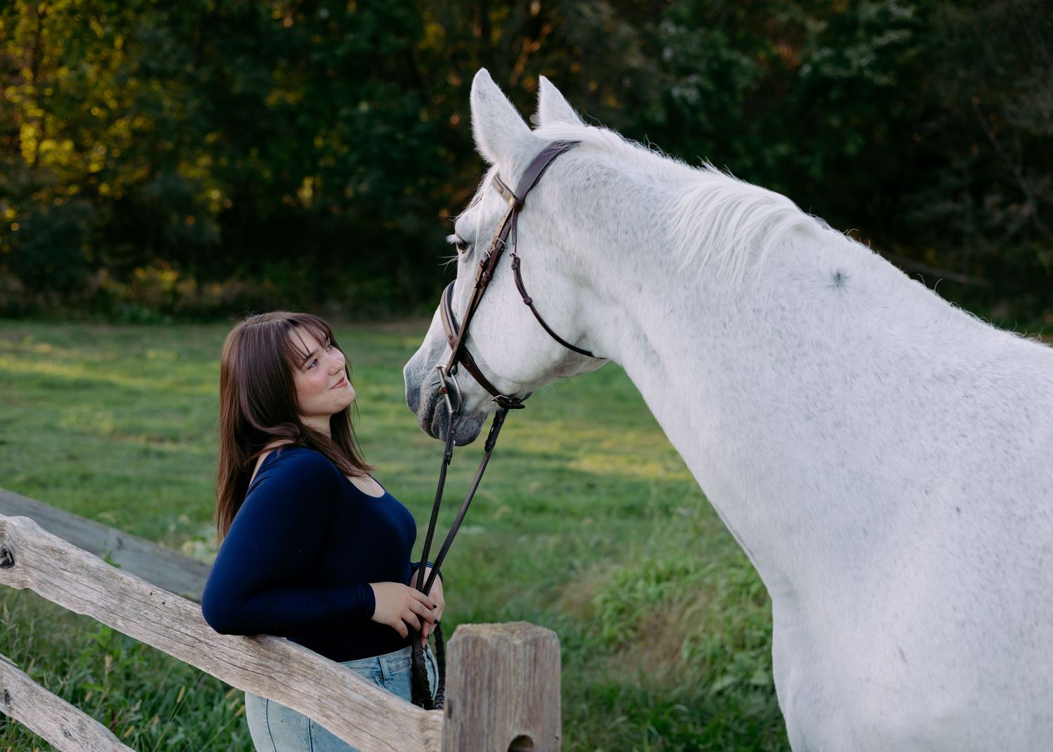 Young woman with long brown hair stands beside a wooden fence, gazing up at a gray horse in a bridle on a green grassy field.
