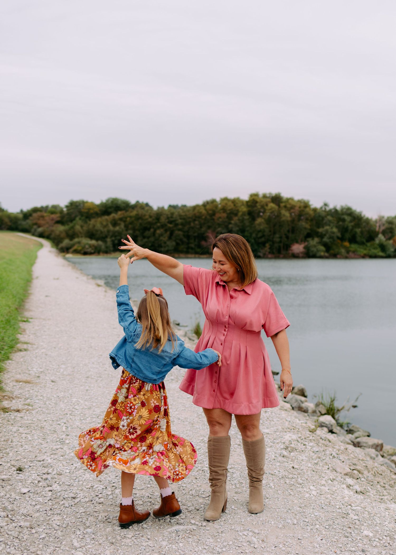 Smiling woman in a pink dress and tall boots twirls a little girl in a floral skirt and denim jacket on a gravel path beside a lake.