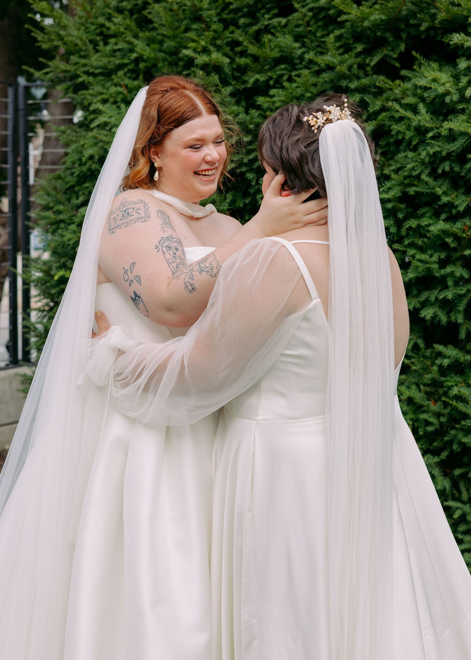 Two brides in white gowns and long veils embrace in an emotional moment outdoors, one smiling with tattooed arms, before a green hedge.