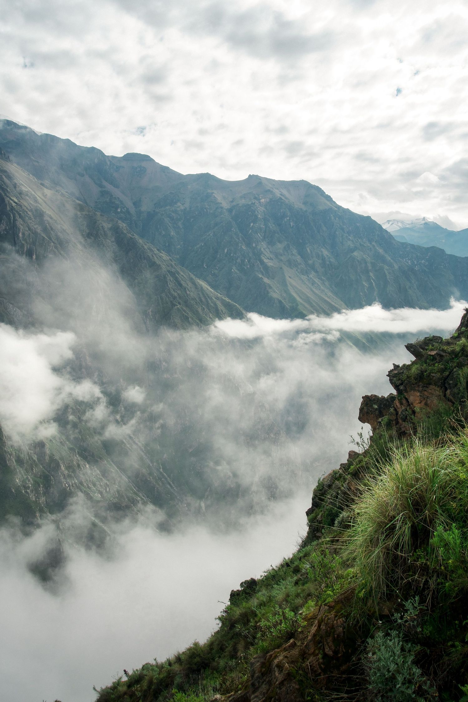 Misty mountain valley with dramatic clouds rolling over steep green slopes and rugged Andean peaks in the background.