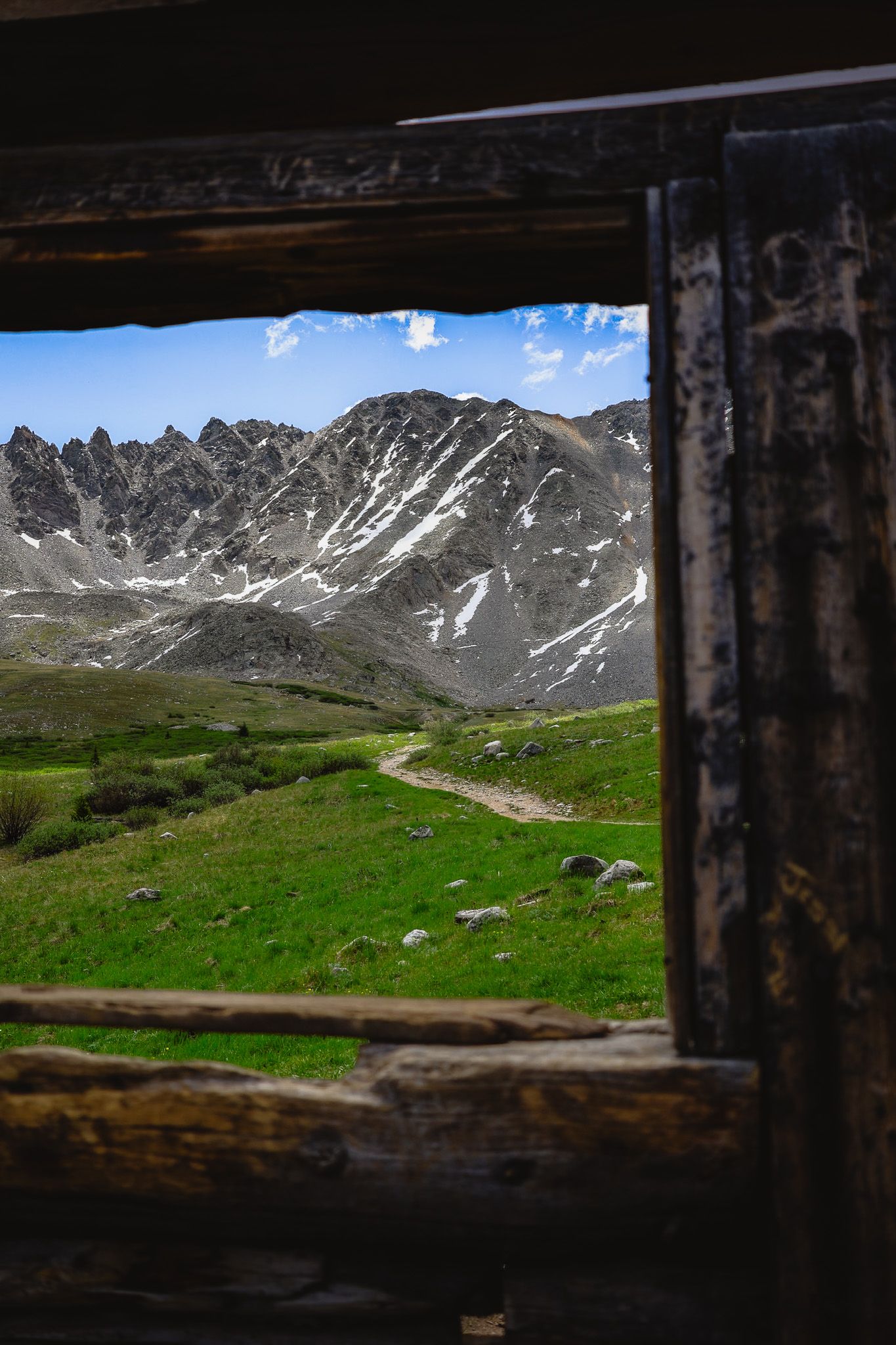 View through rustic wooden window frame of snow-capped mountain peaks, green alpine meadow, and winding trail under blue sky.