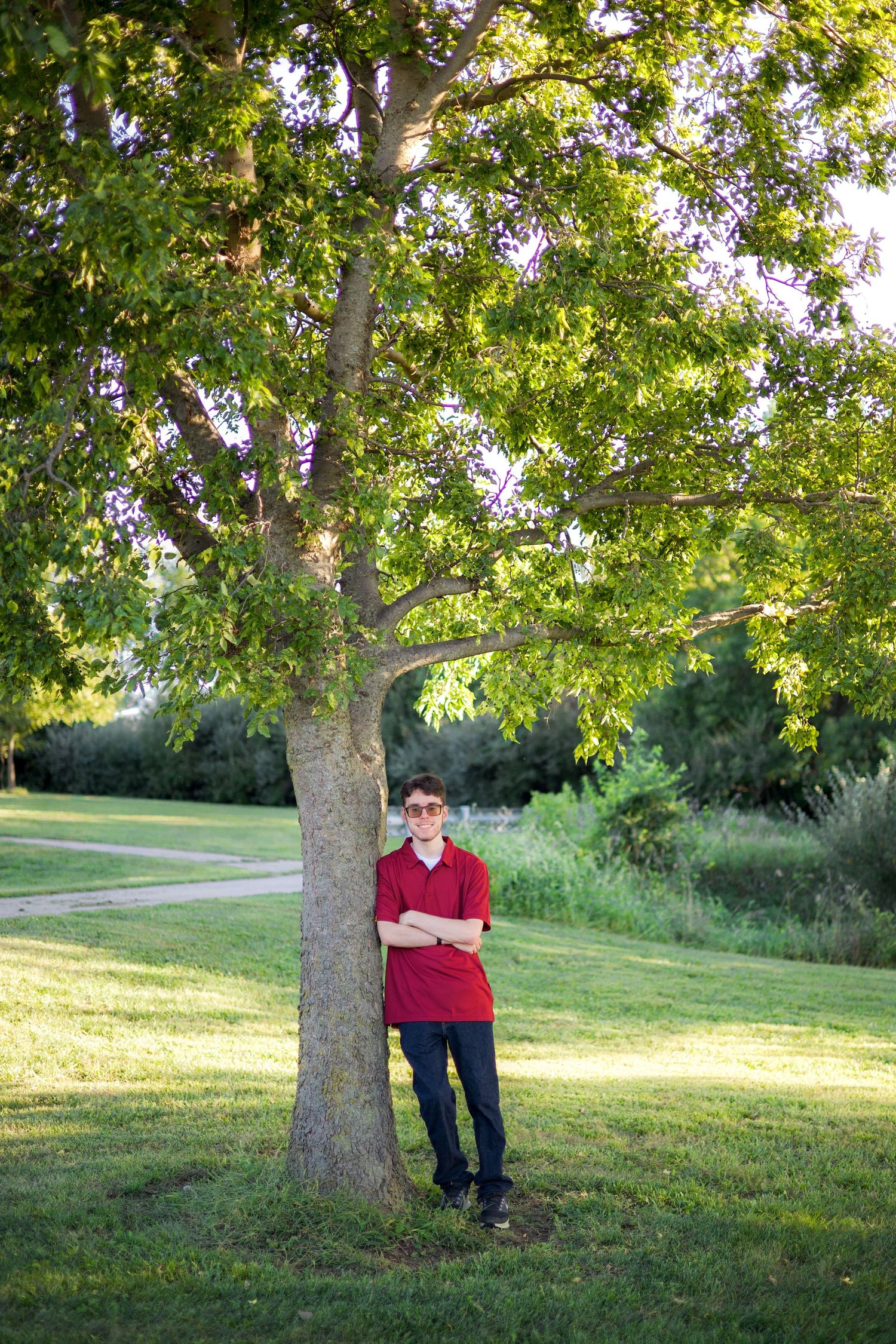 Teen boy in red polo and sunglasses leaning against a tree in a lush green park on a sunny day.