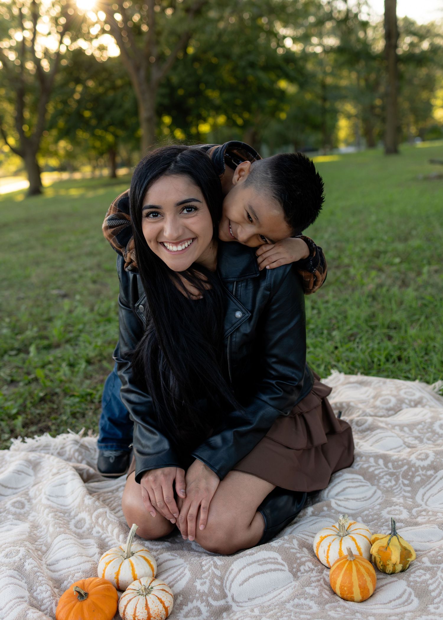 Smiling woman in a black jacket sits on a blanket with small pumpkins as a child hugs her from behind in a sunlit park.