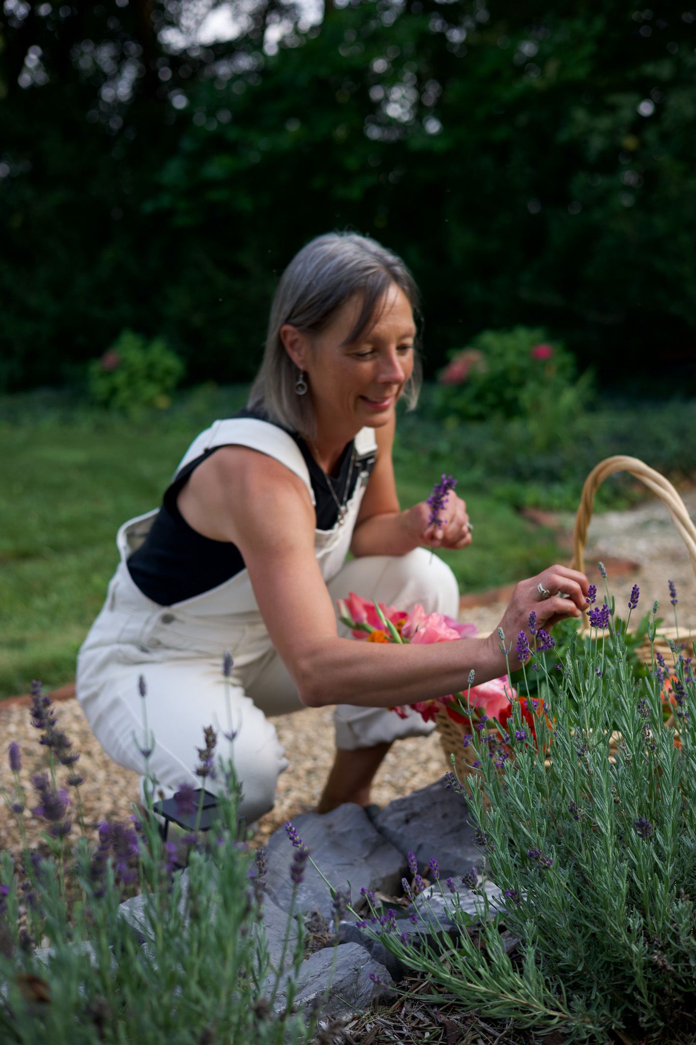 Woman smiling while picking lavender and pink flowers into a wicker basket in a lush garden.