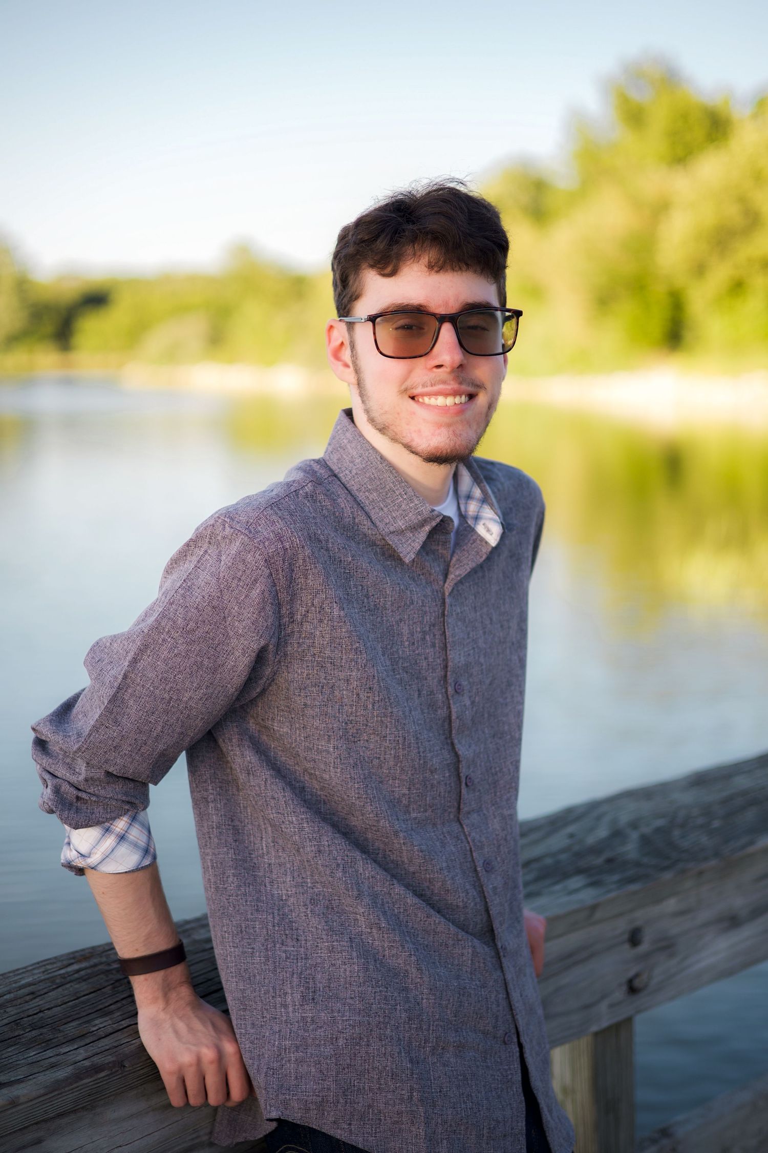 Young man in sunglasses and gray shirt smiling on wooden dock by a calm lake with green trees.