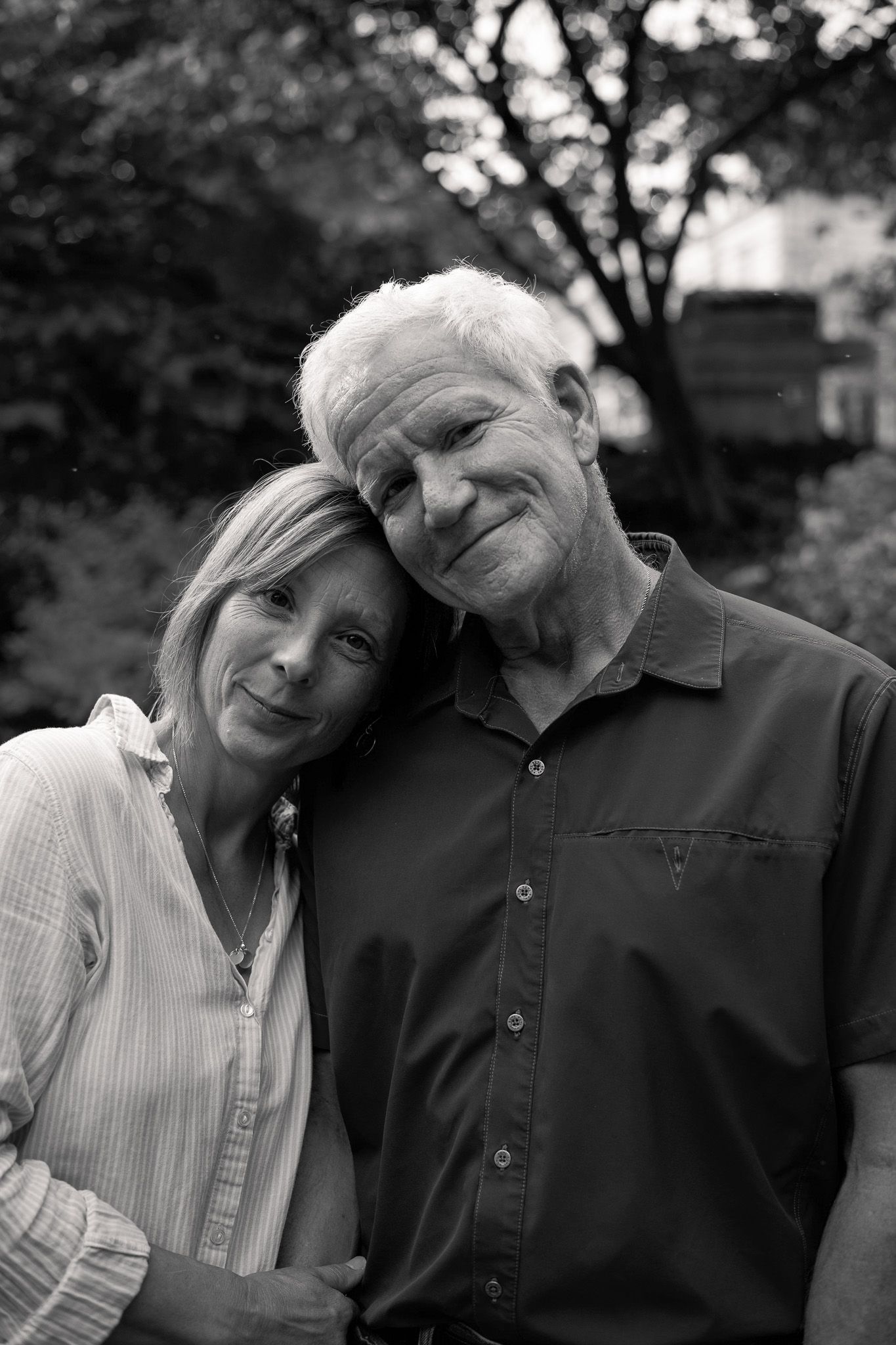 Black and white portrait of a smiling senior couple embracing outdoors with trees in the background.