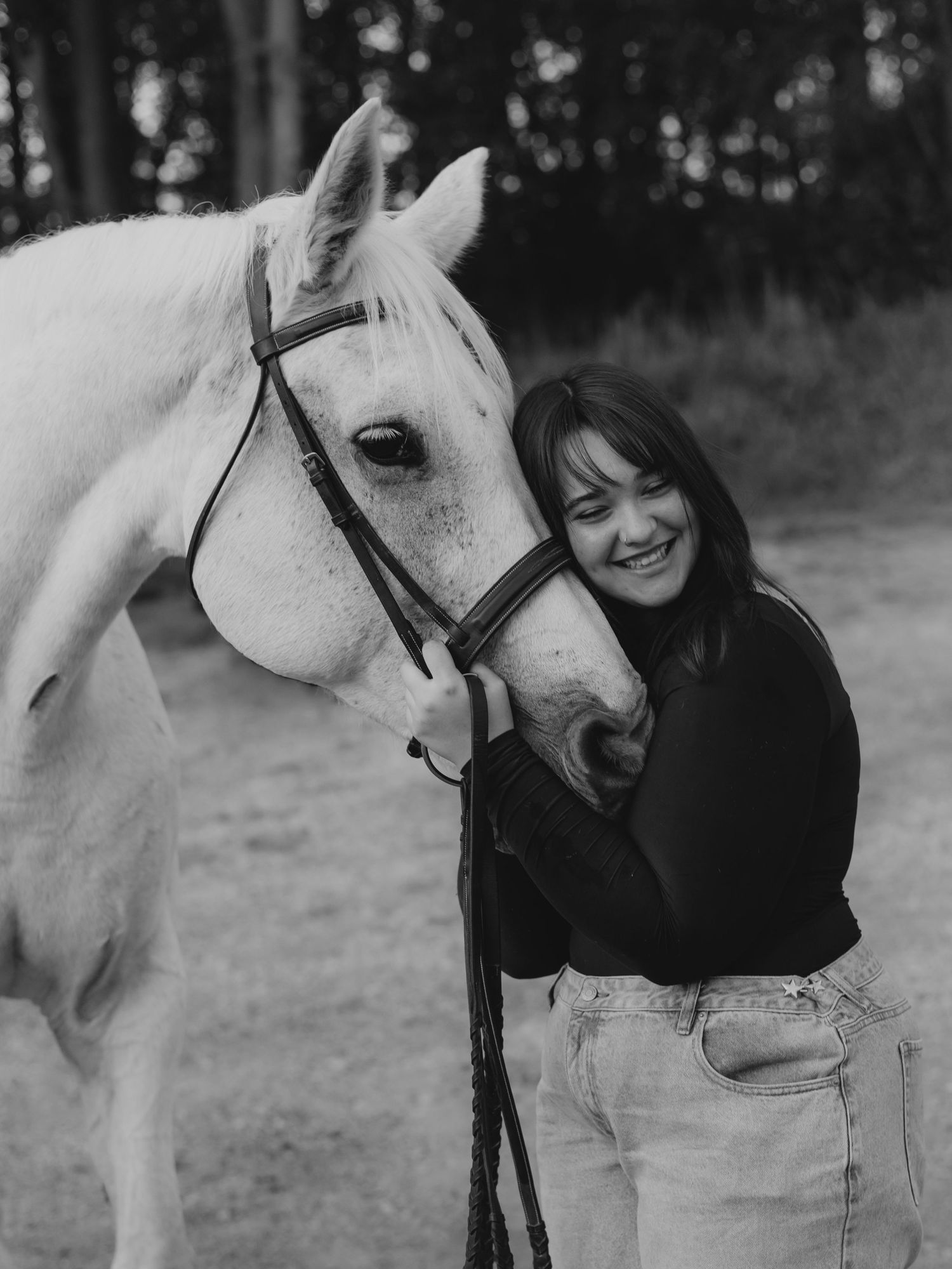 Smiling woman in a dark sweater and jeans hugs a white horse wearing a bridle in a black and white outdoor portrait.