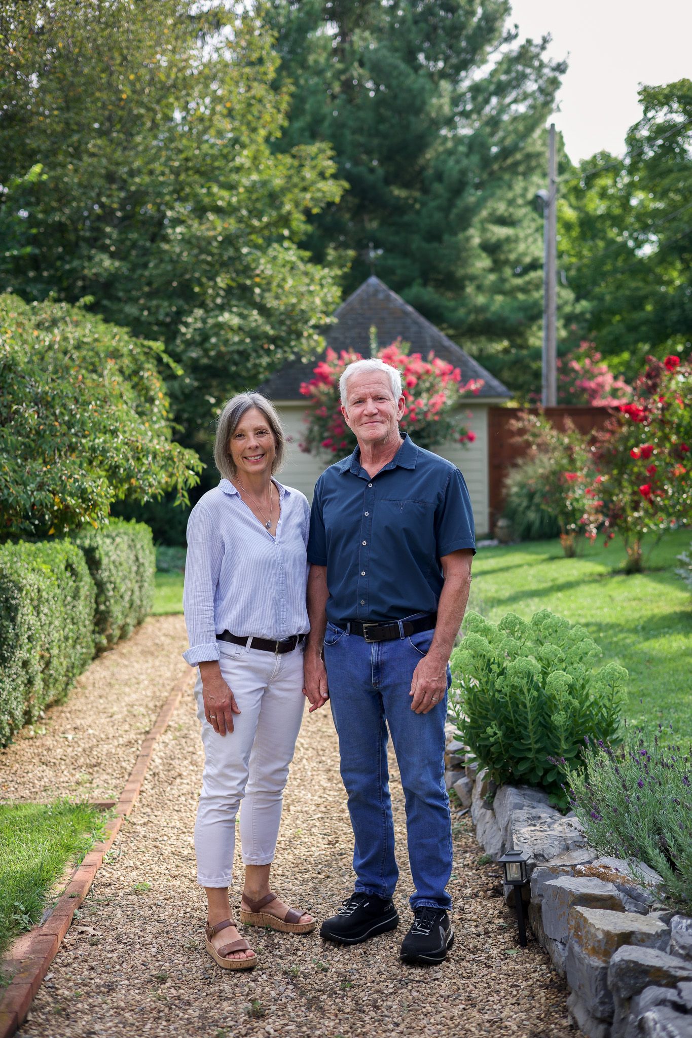 Mature couple smiling and standing together on a gravel garden path with blooming roses behind them.
