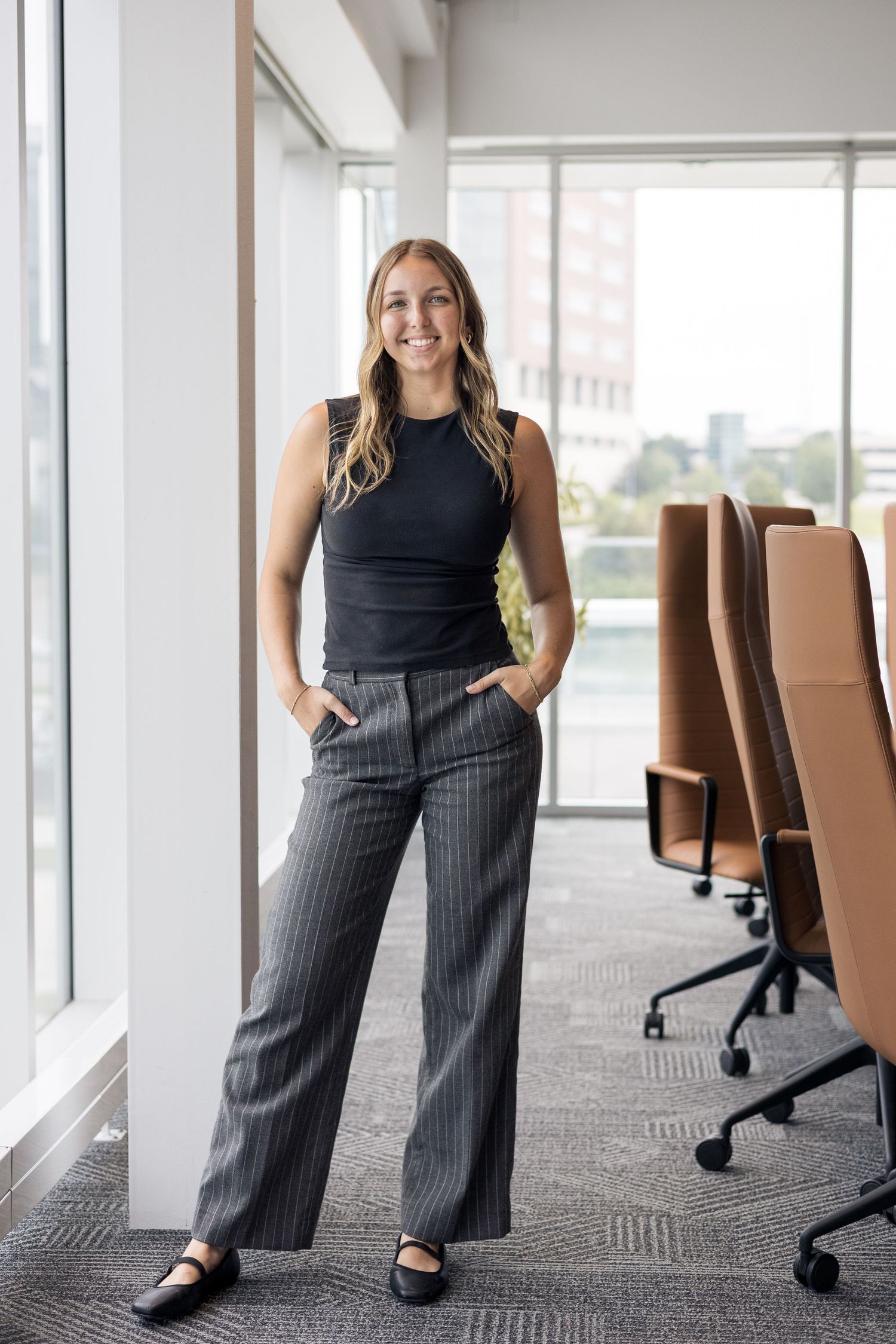 Smiling blonde professional woman in pinstripe trousers standing by office windows in a conference room.