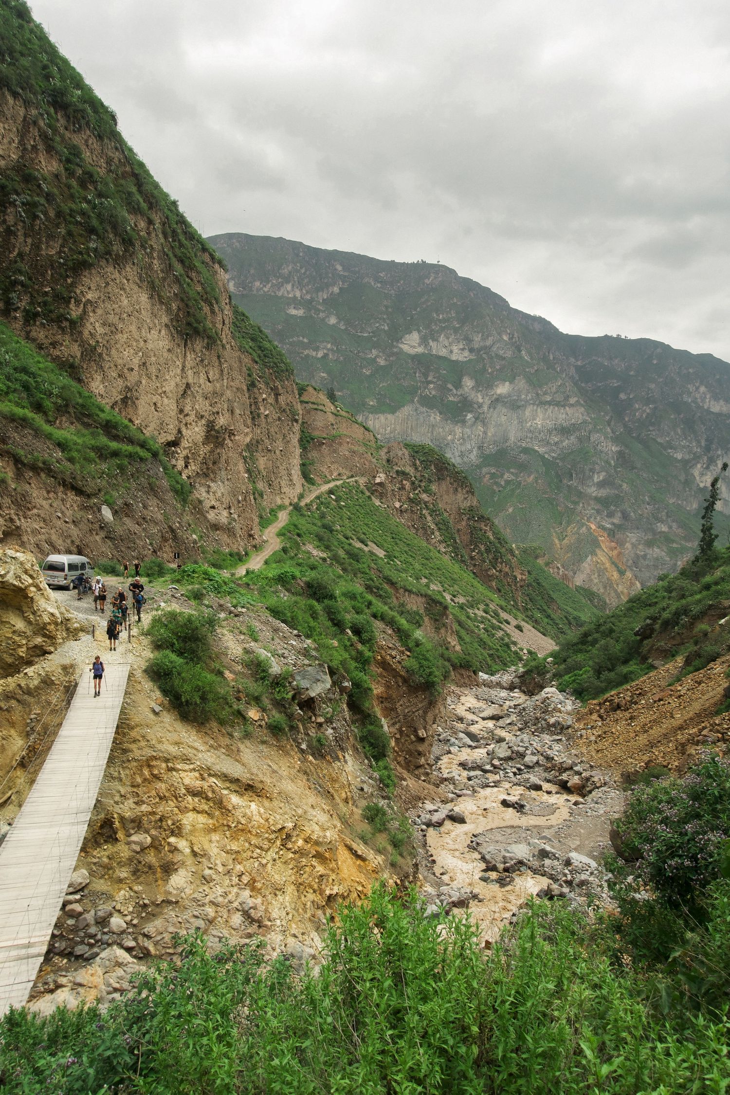 Narrow mountain road with hikers and a bus winding through a steep rocky gorge with lush green vegetation.