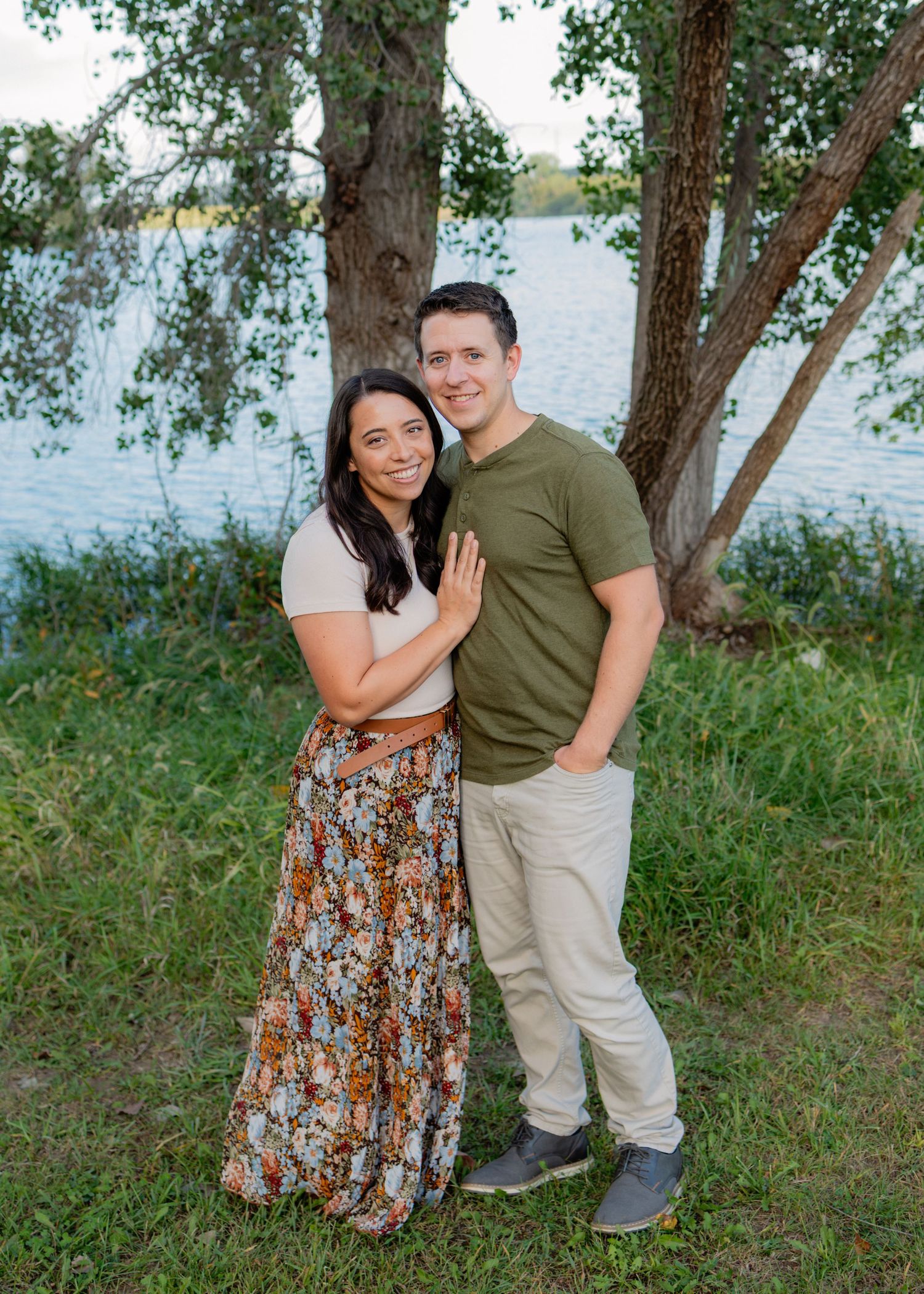 A smiling couple embraces on a grassy lakeside, she in a floral maxi skirt and he in an olive t-shirt, with trees and water behind them.