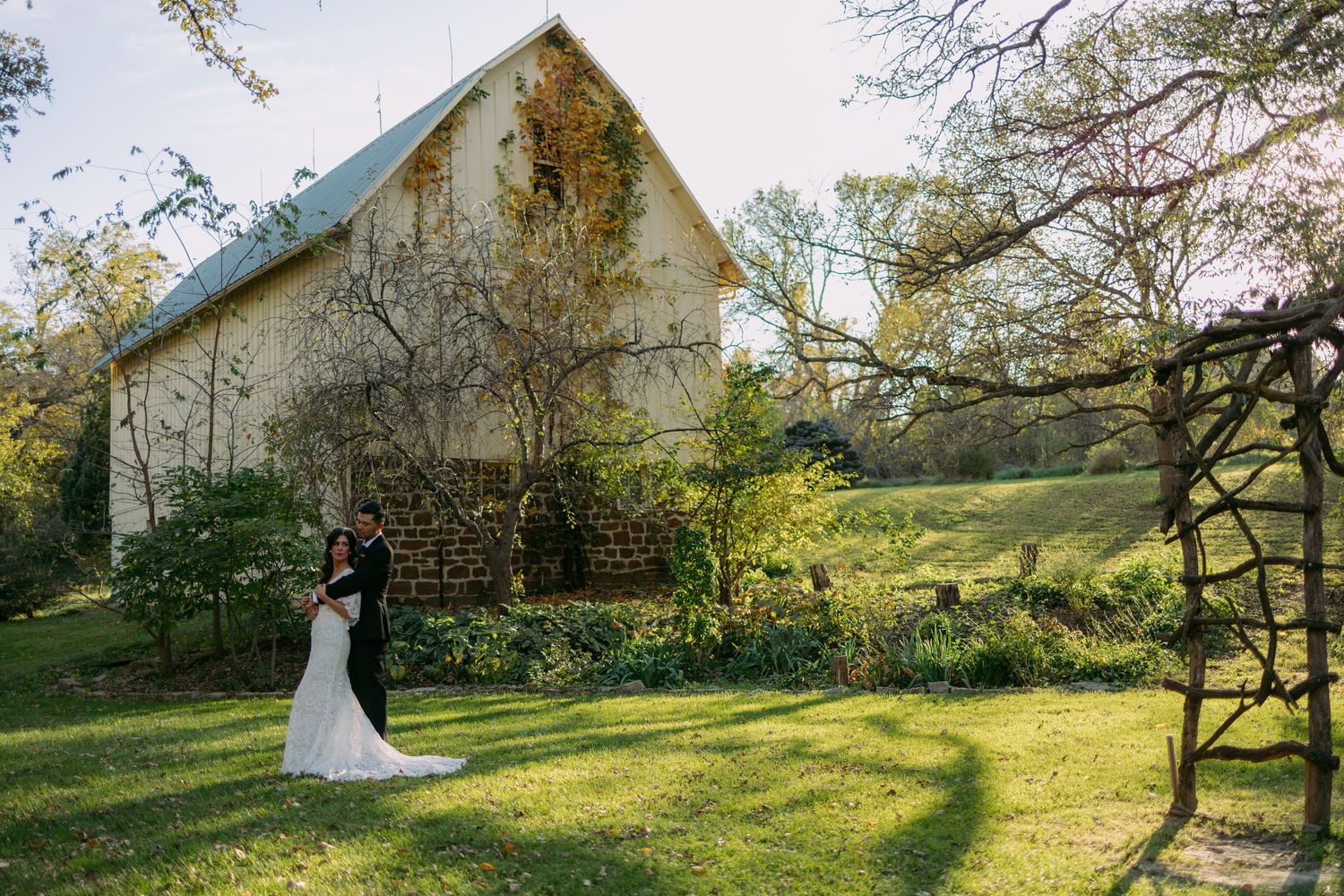 Bride in a white gown and groom in a dark suit embrace on a sunlit lawn beside a rustic stone barn wedding venue surrounded by trees.