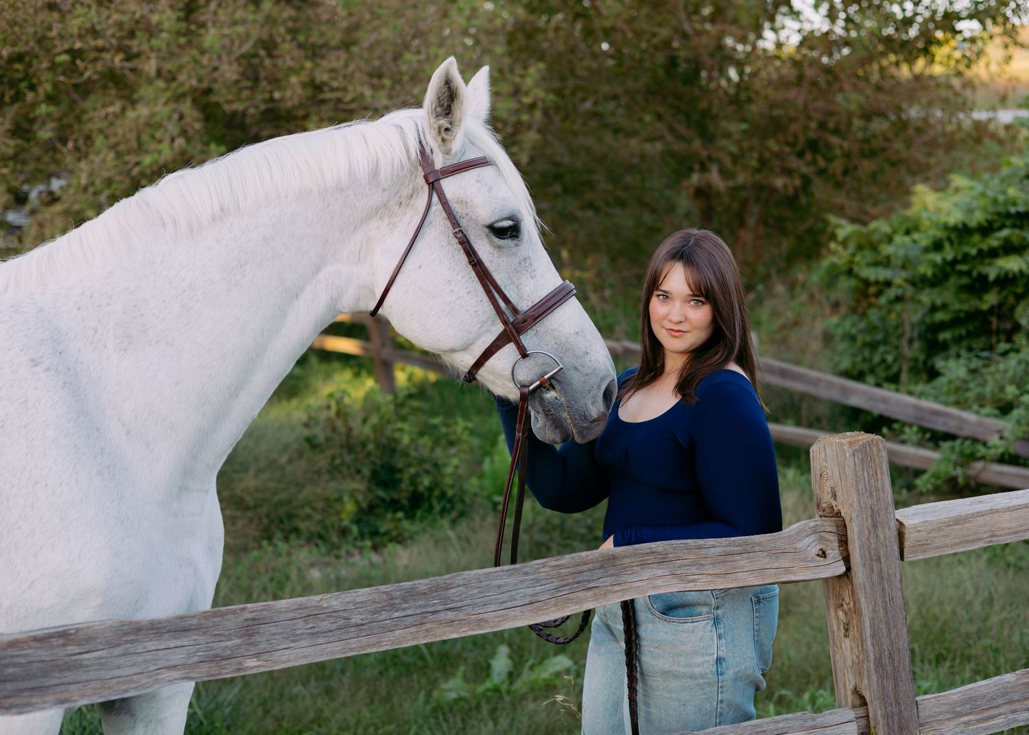 Smiling woman in a navy sweater and jeans stands beside a gray horse in a bridle at a wooden fence with green trees behind them.