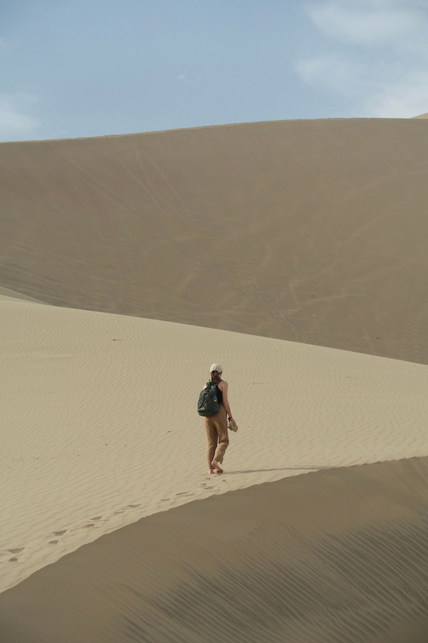 A lone hiker with a backpack walks barefoot across vast golden sand dunes under a pale blue sky.