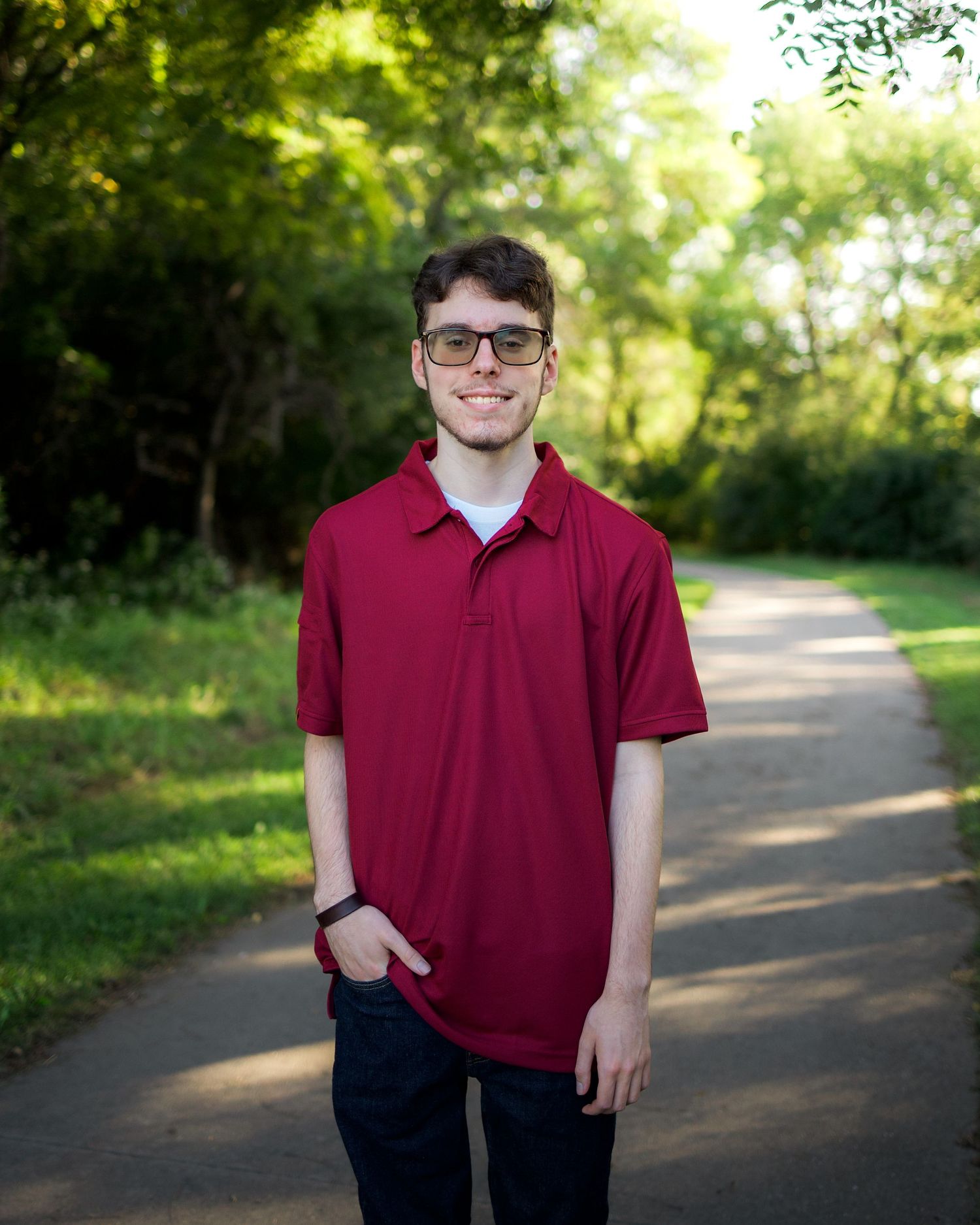 Young man in red polo shirt and glasses smiling on a tree-lined path outdoors.