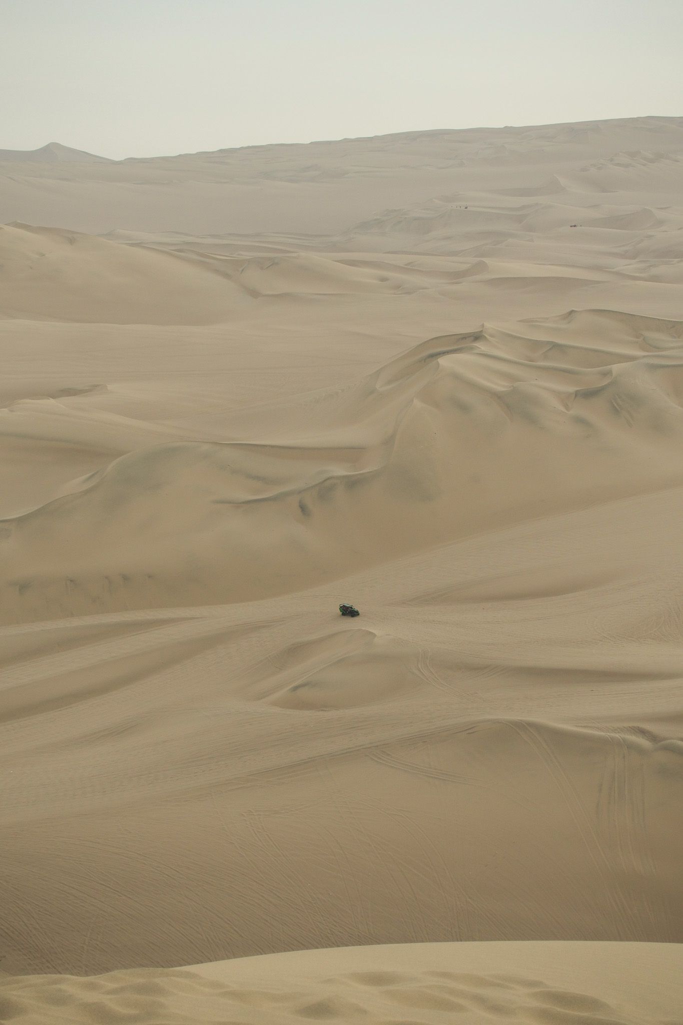 Vast golden sand dunes stretch endlessly in a desert landscape with a tiny vehicle navigating the remote terrain.