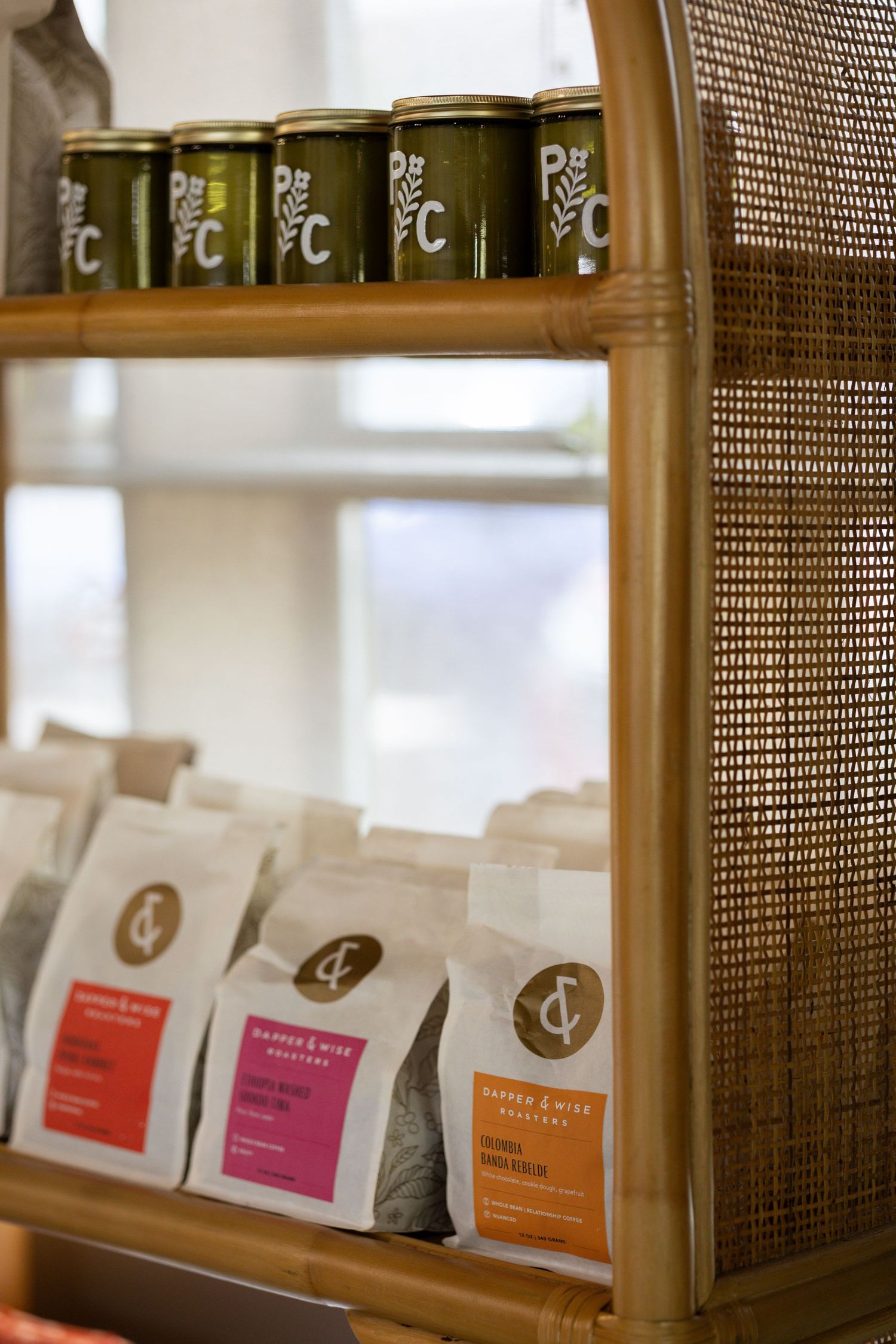 Rattan display shelf with green canned drinks on top and white coffee bags with colorful labels on lower shelf.