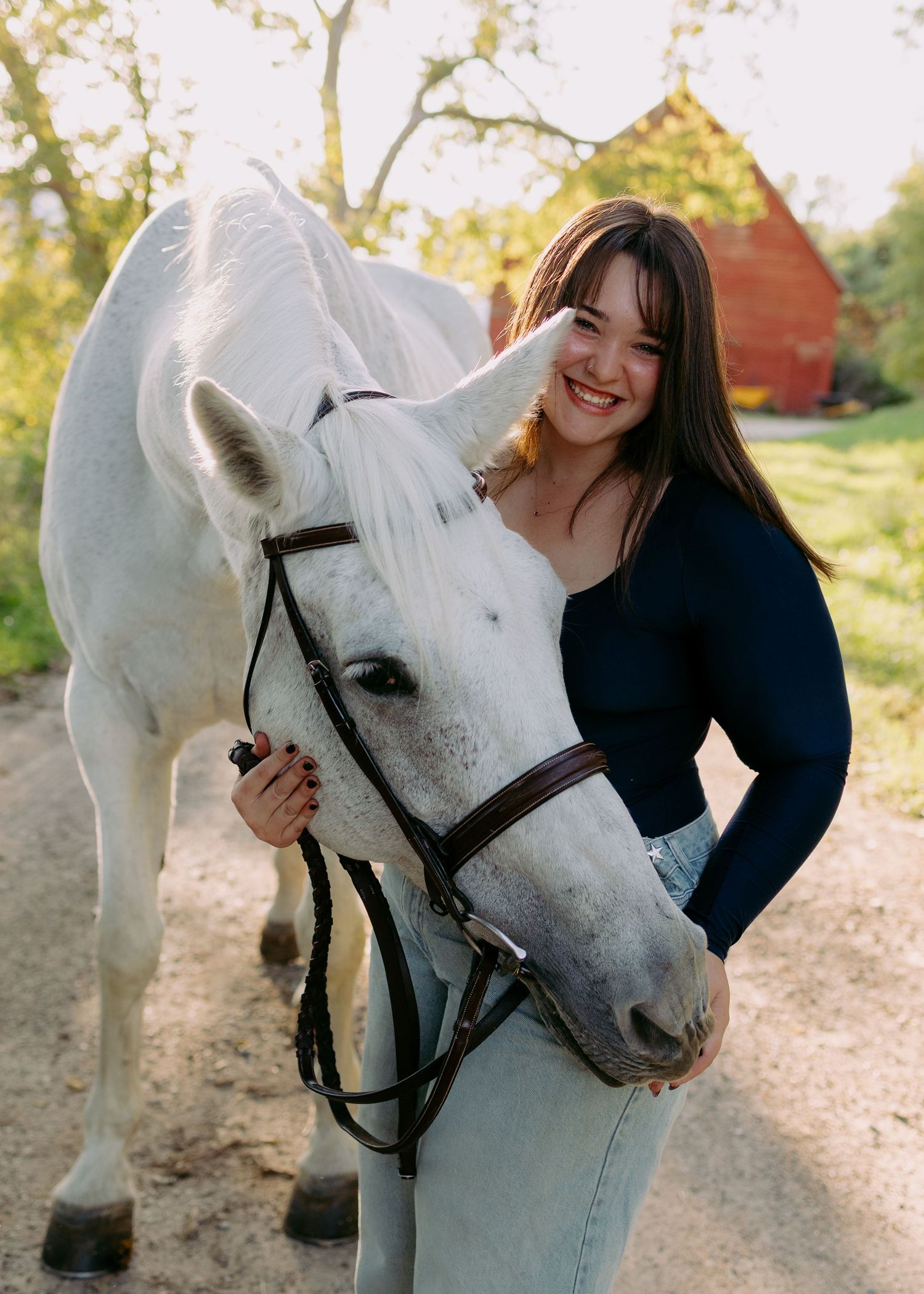 Smiling young woman with long brown hair hugging a gray horse wearing a bridle on a dirt path near a red barn.