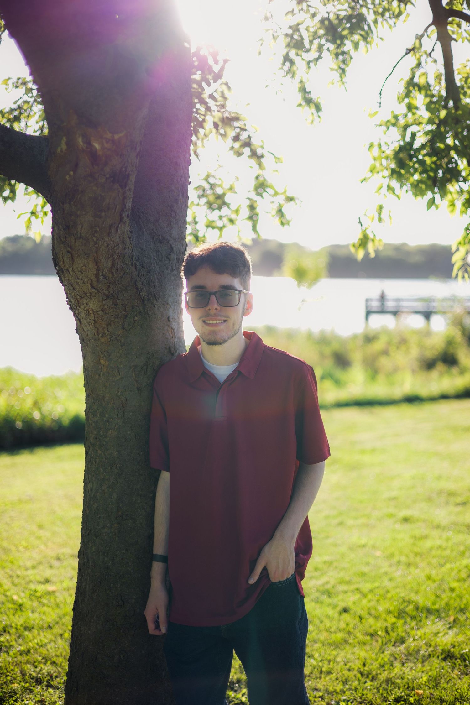 Young man in red shirt and sunglasses smiling beside a tree in a sunny park near a lake.