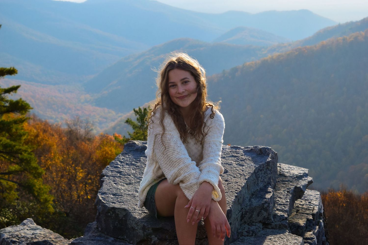 Young woman sitting on rocky mountain overlook with colorful autumn forest valley view behind her.