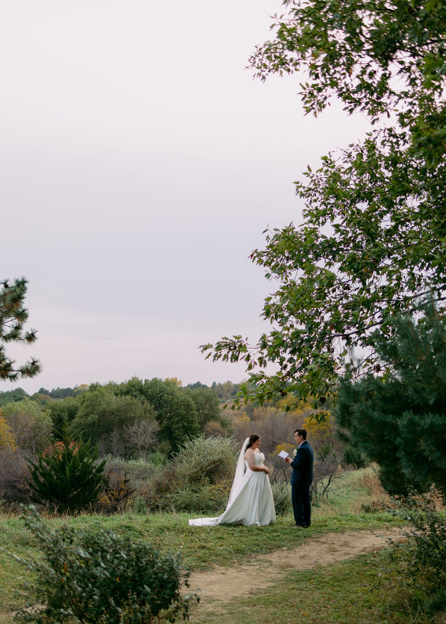 Bride in a white gown and groom in a dark suit stand facing each other on a grassy path surrounded by trees at dusk.