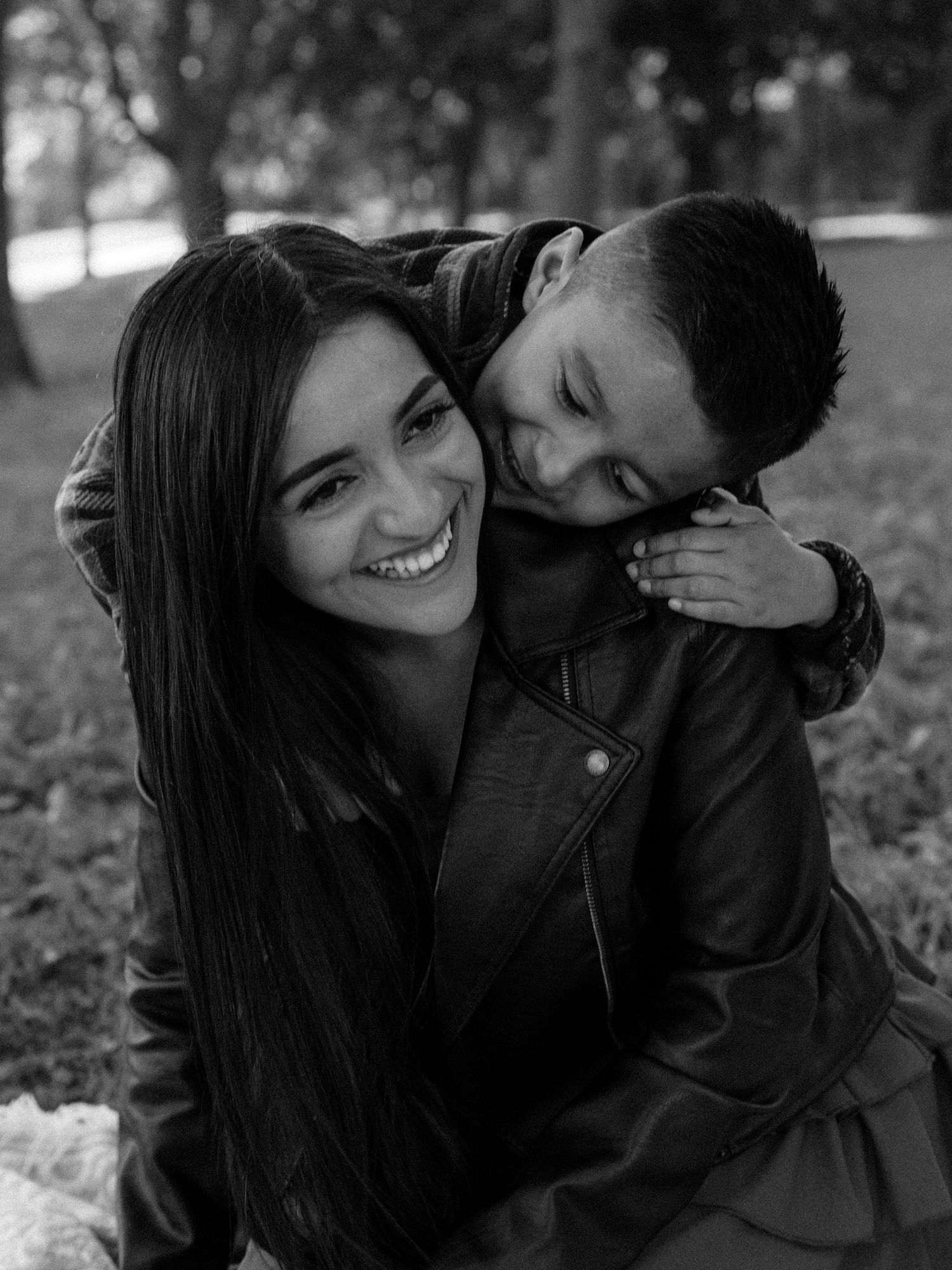 Smiling woman with long dark hair in a leather jacket sits on grass as a young boy hugs her from behind in a black-and-white photo.