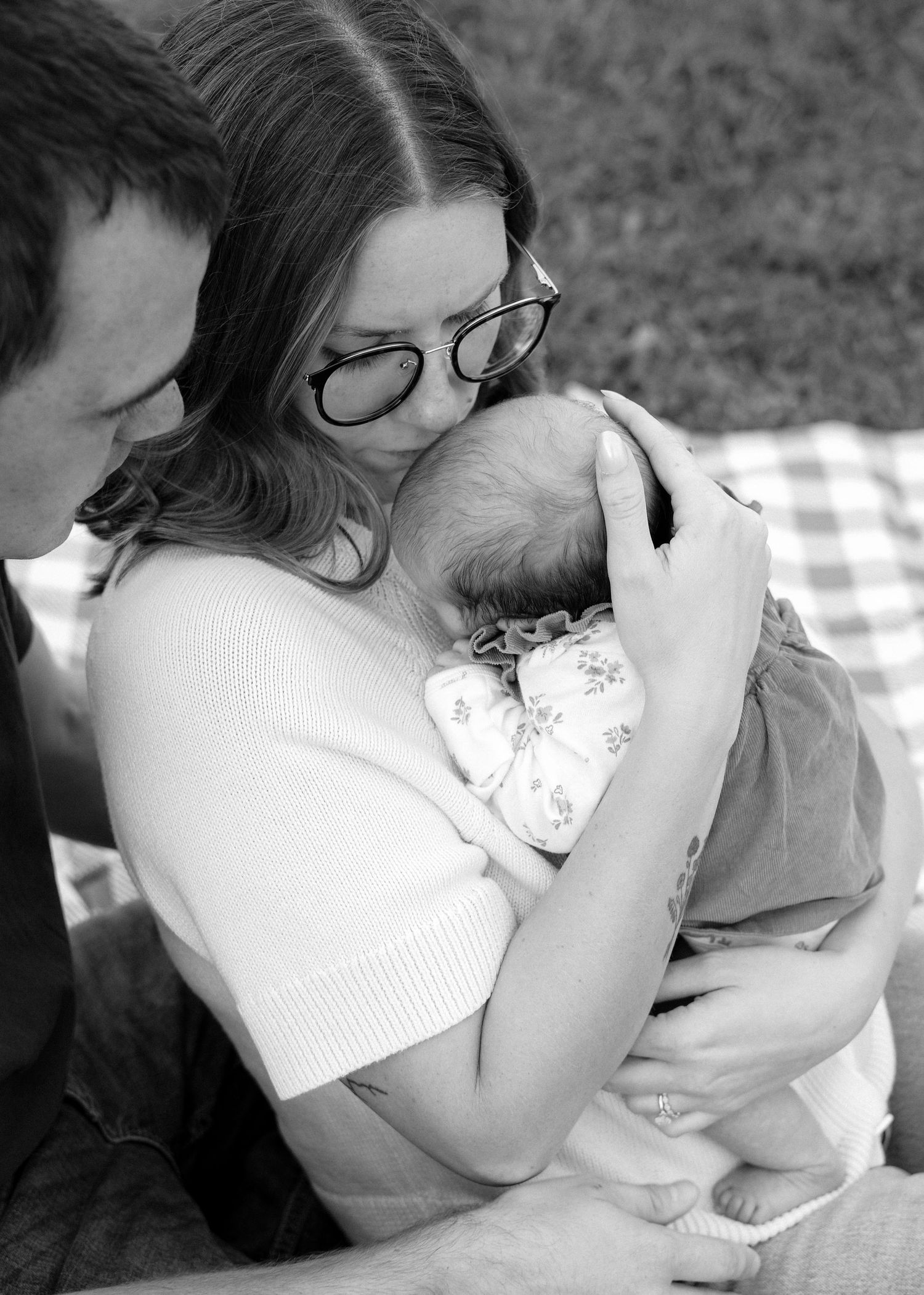 Black and white photo of a mother kissing her newborn baby while a man looks on during an outdoor family session.
