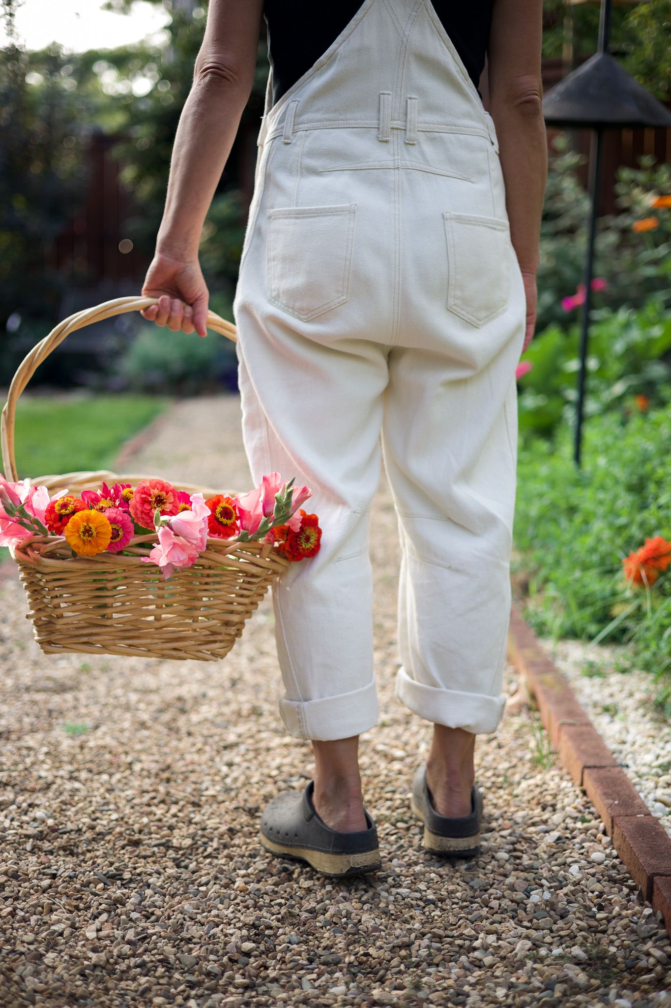 Person in white overalls carrying a wicker basket filled with colorful flowers walking along a garden gravel path.