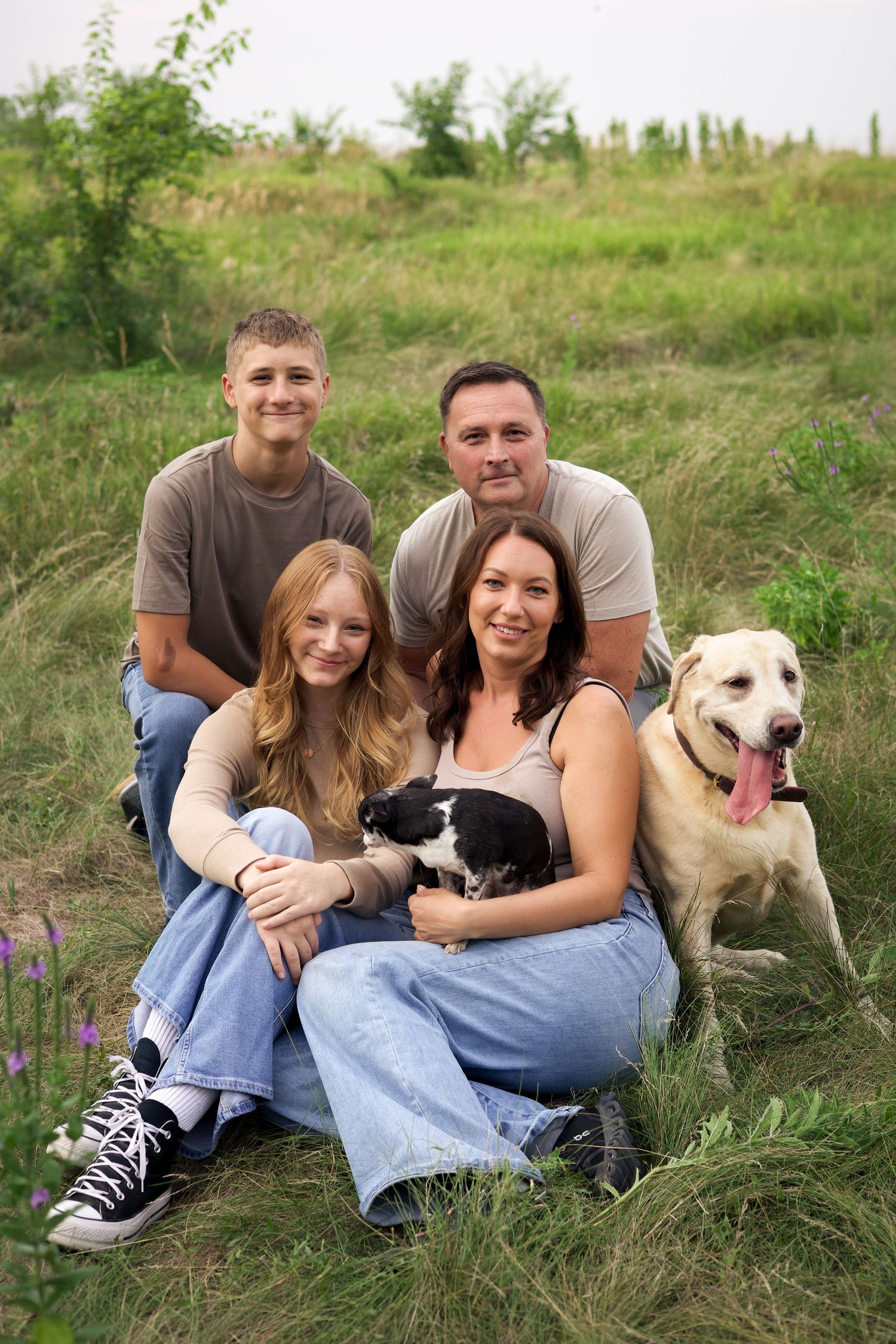 Family of four with two dogs sitting in a green field, smiling together for an outdoor portrait.