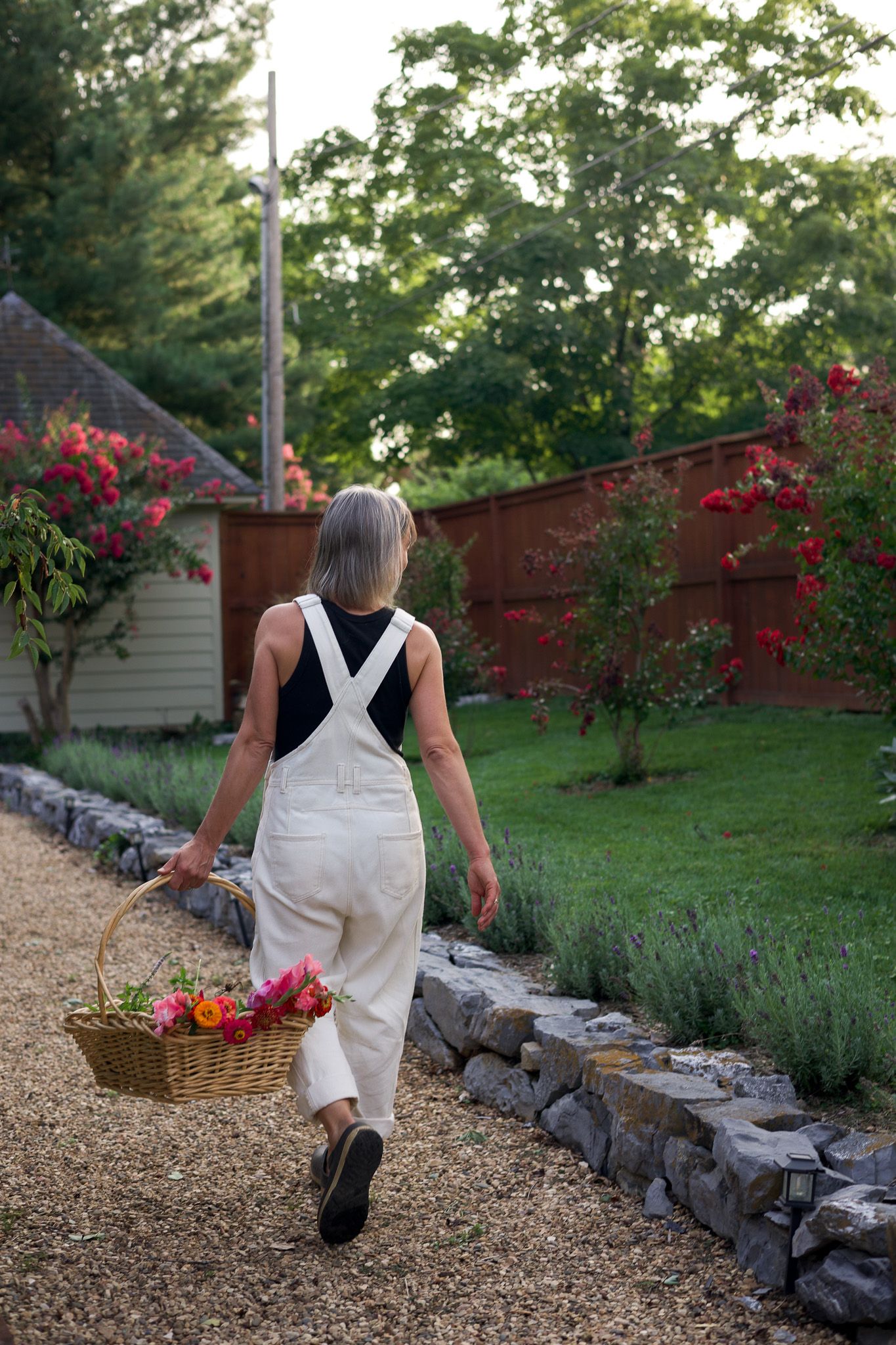 Woman in white overalls carrying a wicker basket of flowers through a lush garden path lined with lavender.