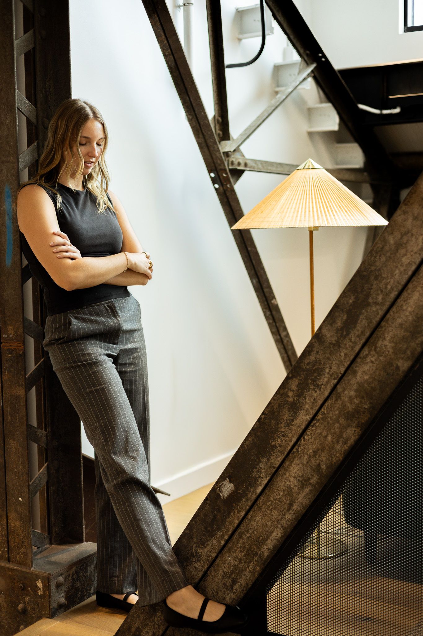 Woman in gray pinstripe pantsuit leaning against industrial staircase beside a conical bamboo floor lamp.