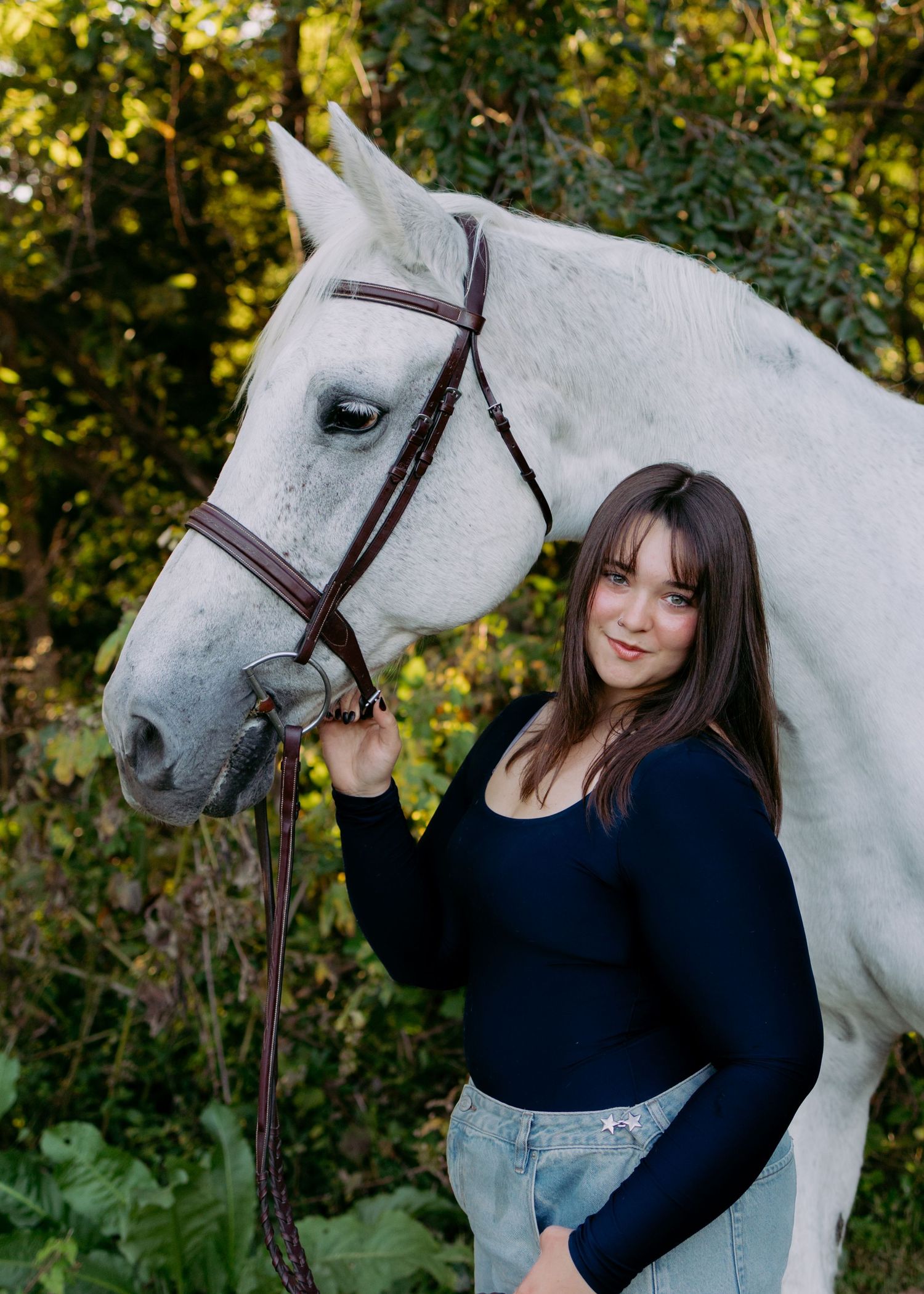 Young woman with long brown hair in a navy top and jeans standing beside a gray horse in a leather bridle among green foliage.