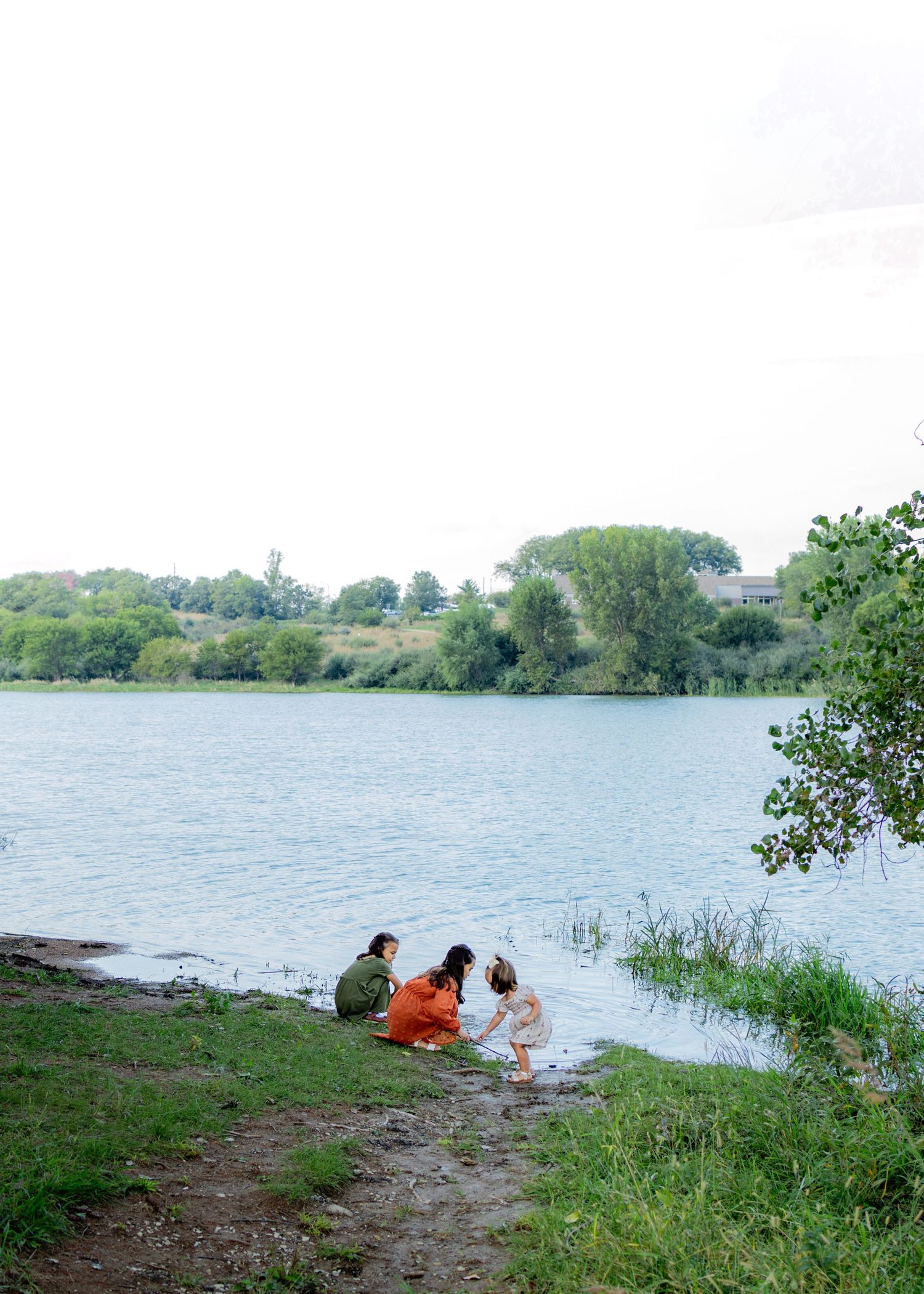 Family sitting by a calm lake shore surrounded by lush green trees and grass on a cloudy day.