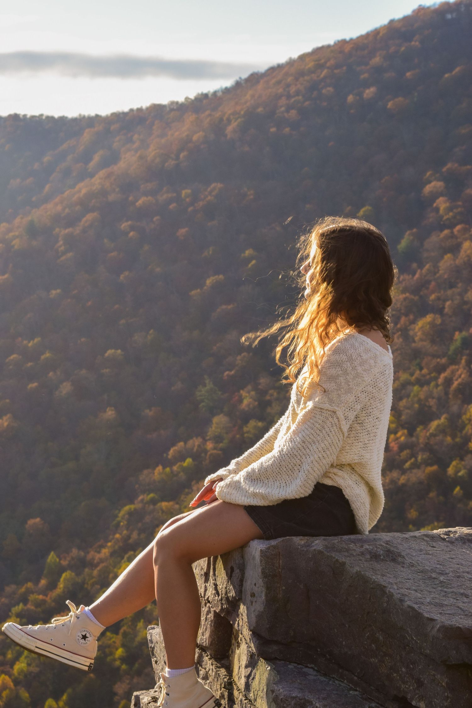 Young woman in cream sweater and Converse sits on rocky ledge overlooking autumn mountain forest at golden hour.