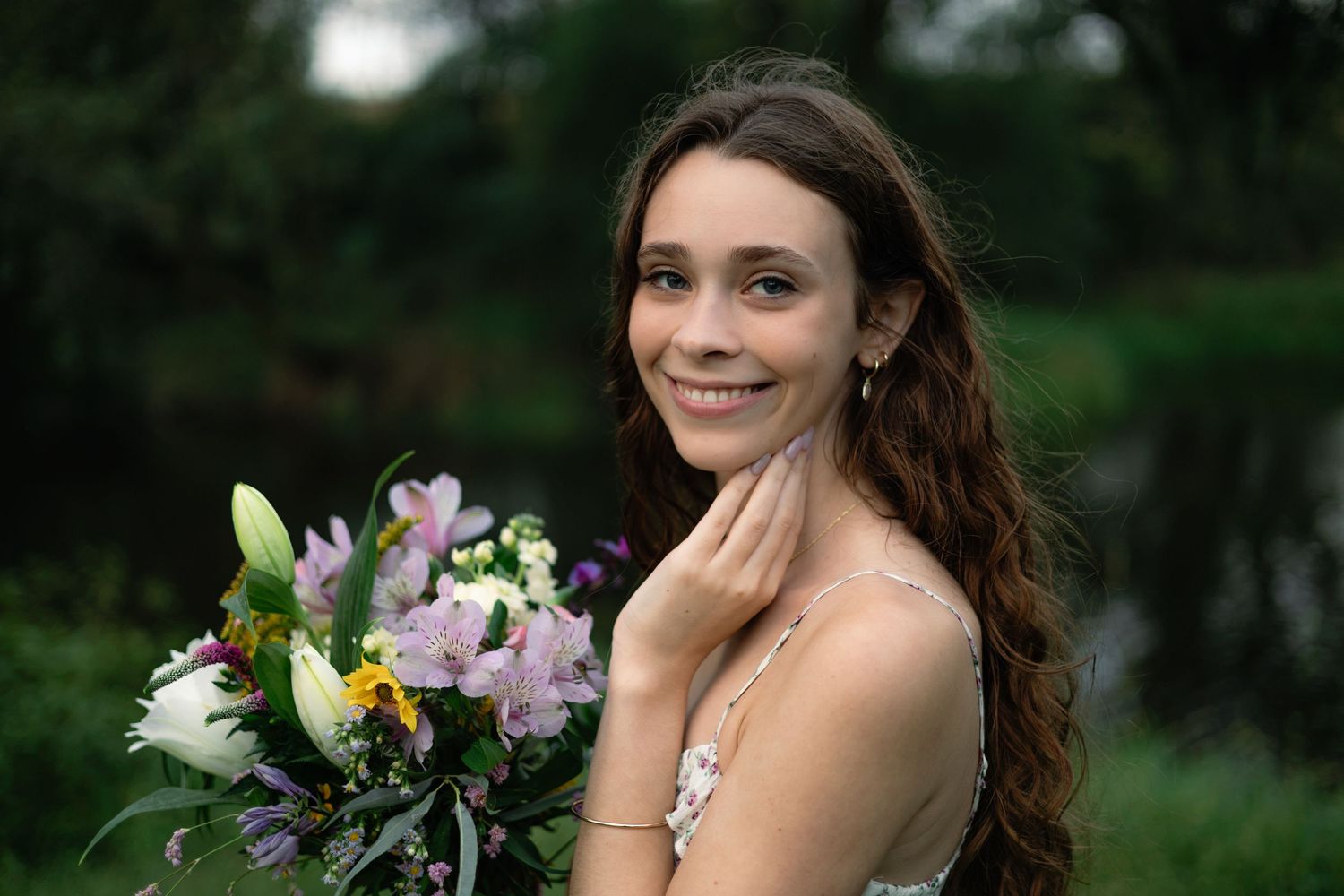 Young bride smiling with colorful flower bouquet outdoors, wearing elegant dress and earrings with green trees background.