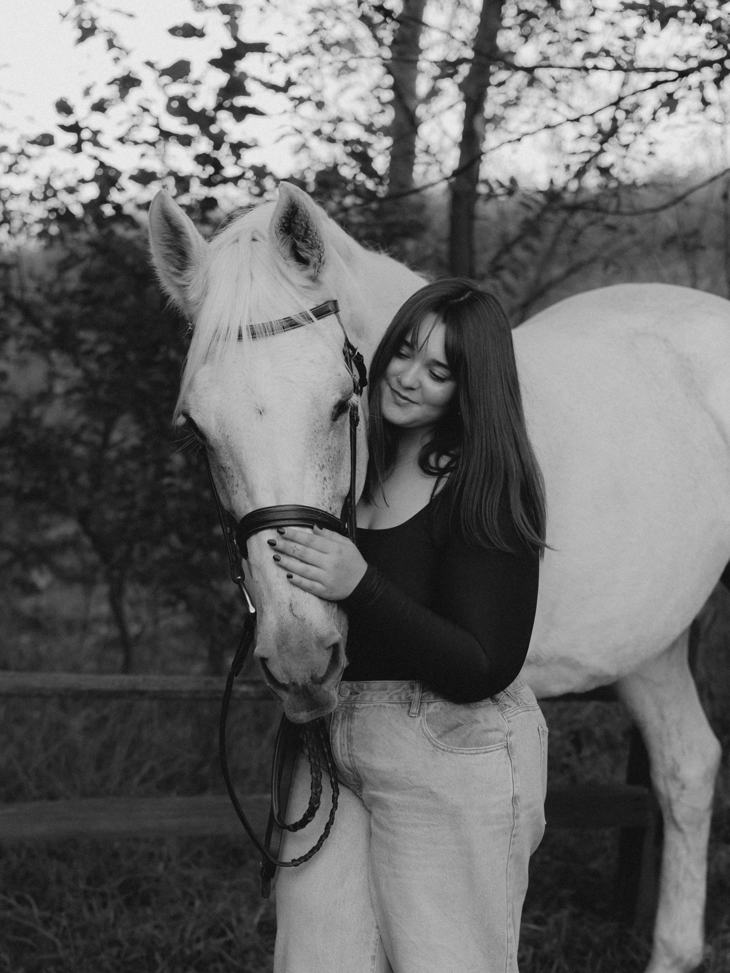 Black and white portrait of a smiling young woman with long dark hair gently embracing a light-colored horse in a wooded pasture.