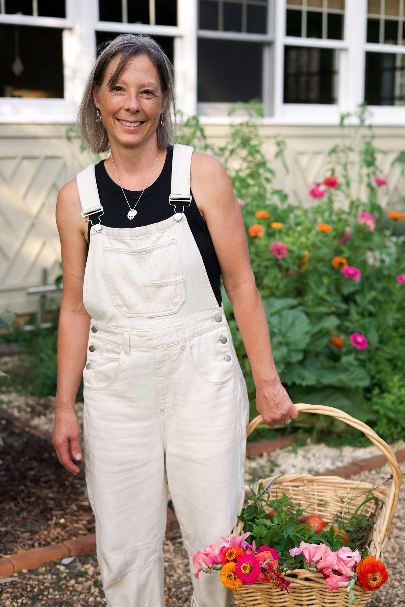 Smiling woman in white overalls holding a wicker basket of colorful flowers in a garden.